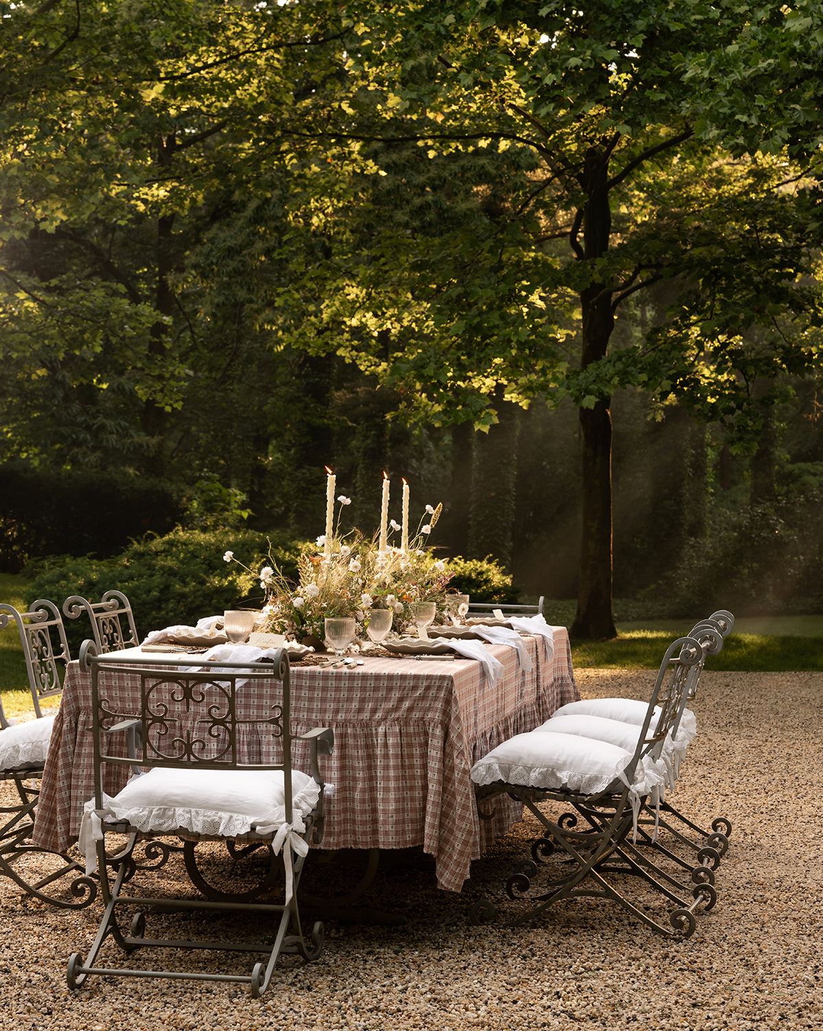 Certainly! Here’s your sentence with the exact string McGee & Co. blended at the end: An elegant outdoor table with a checked cloth and flowers is surrounded by six iron chairs, each topped with a McGee & Co. x Loeffler Randall Delilah Broderie Trim Seat Cushion, all beneath sun-dappled trees on gravel by McGee & Co.