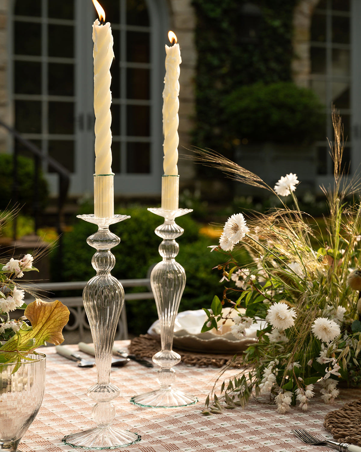 Two tall ivory candles in McGee & Co. x Loeffler Randall Effie Taper Holders sit on a checkered tablecloth, surrounded by white and beige dried flowers and greenery, with a blurred garden and building in the background, by McGee & Co.