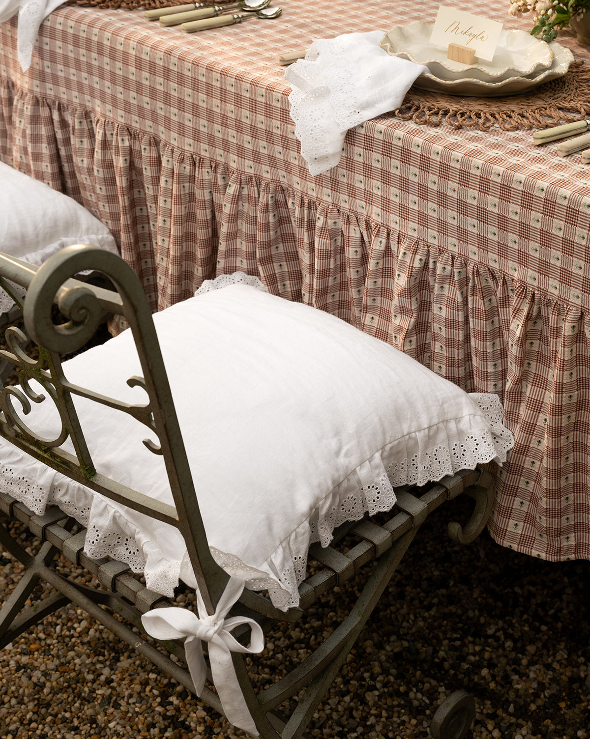 A vintage-style table with a ruffled, plaid tablecloth and arranged cutlery stands on gravel, featuring an ornate metal chair topped with the McGee & Co. x Loeffler Randall Delilah Broderie Trim Seat Cushion, blending modern elegance with timeless charm from McGee & Co.