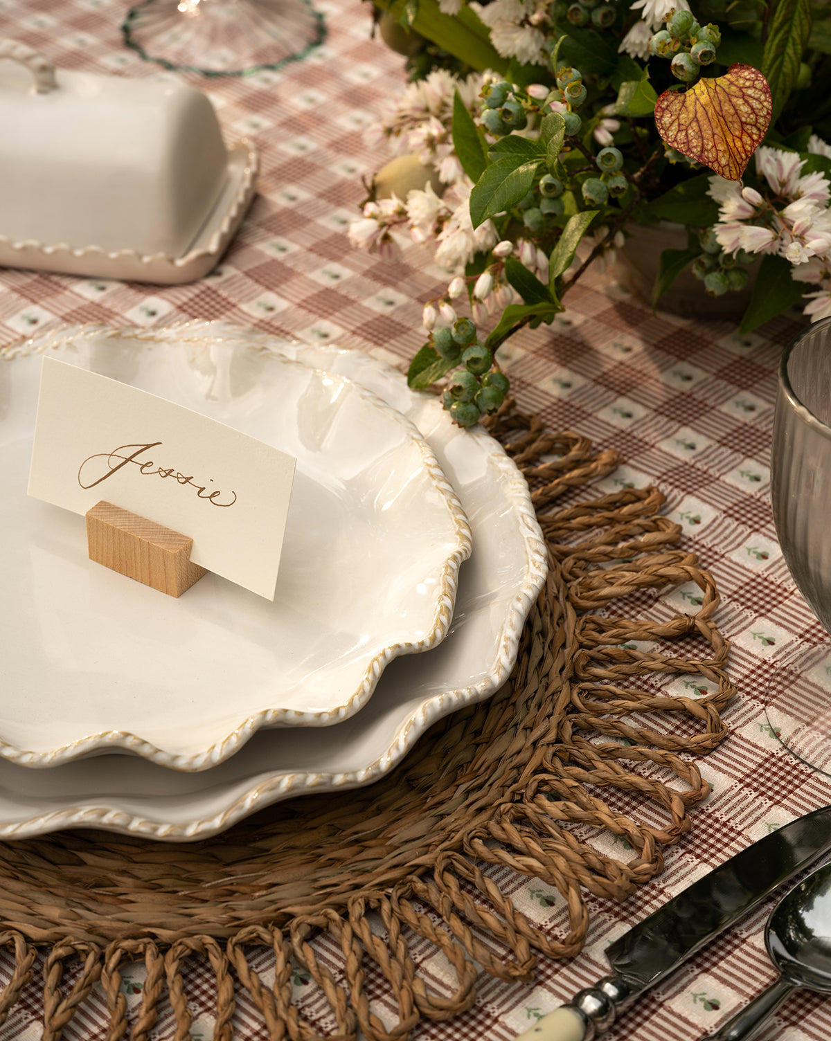 A place setting on a checkered tablecloth features two scalloped white plates, cutlery, a cup, elegant greenery with small white flowers, a handwritten Jessie name card, and the McGee & Co. x Loeffler Randall Elise Seagrass Placemat, by McGee & Co.