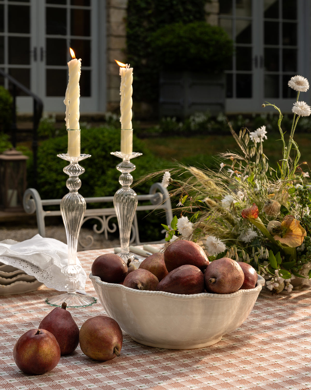 The McGee & Co. x Loeffler Randall Tabitha Serving Bowl, filled with red pears, rests on a checkered tablecloth with two lit candles and wildflowers, set outdoors amid lush garden greenery by McGee & Co.