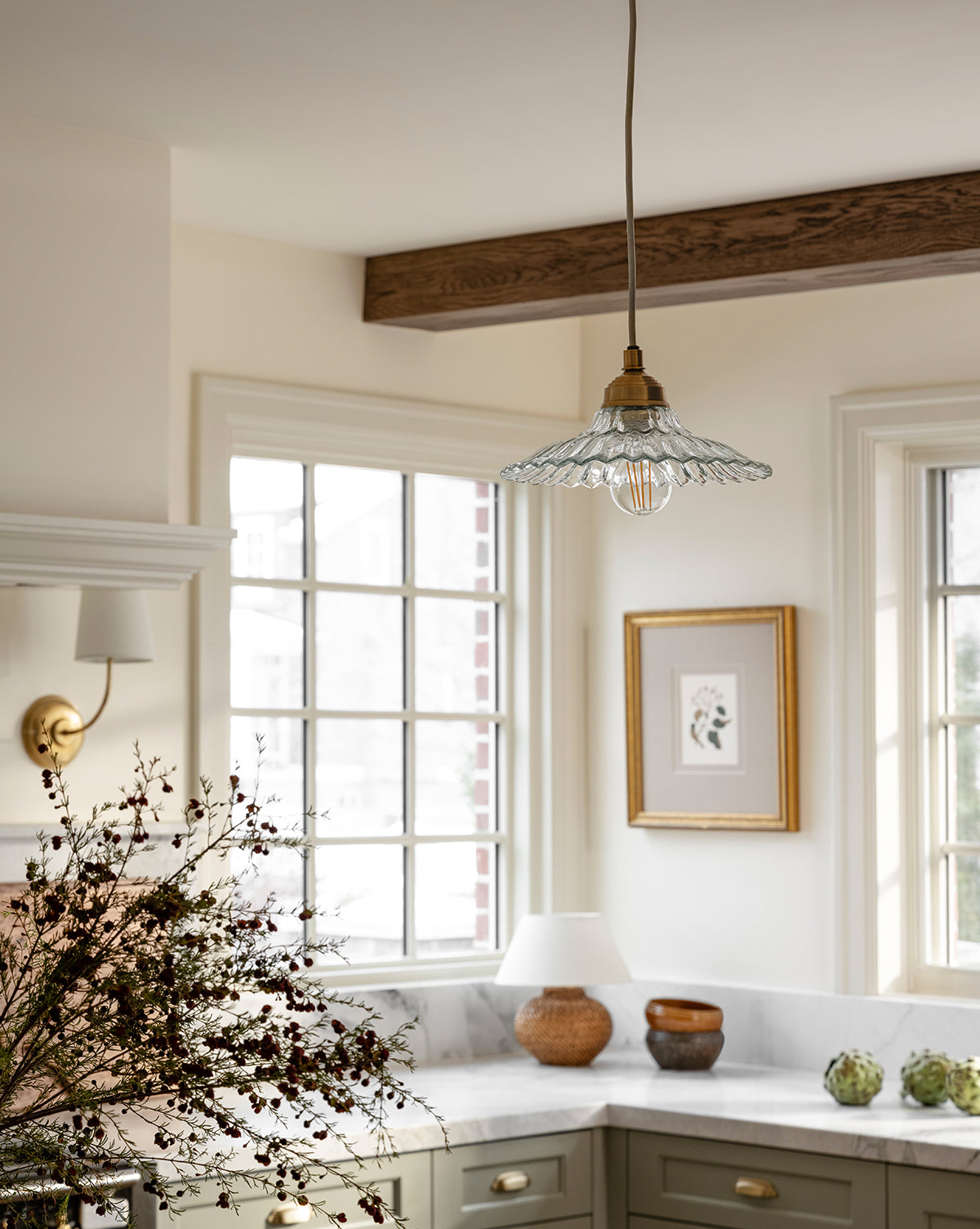 A bright kitchen with a McGee & Co. x Loeffler Randall Maude Glass Pendant, ruffled glass shade, brass hardware, large windows, green cabinets, marble counters, a framed wall picture, and a vase of dried flowers on the countertop by McGee & Co.