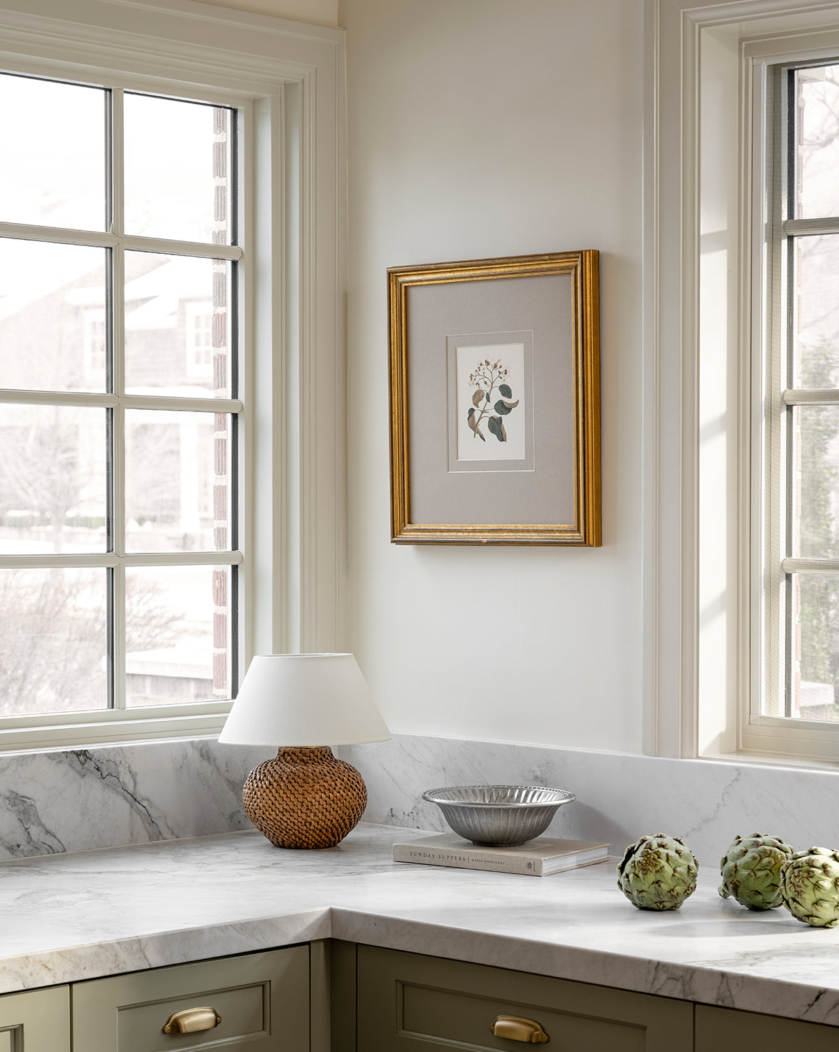 A sunlit kitchen corner features large windows, a marble countertop, the Avedon Cordless Accent Lamp with a white shade, a framed botanical print, a bowl holding two artichokes, and the book The Perfect Kitchen, by McGee & Co.