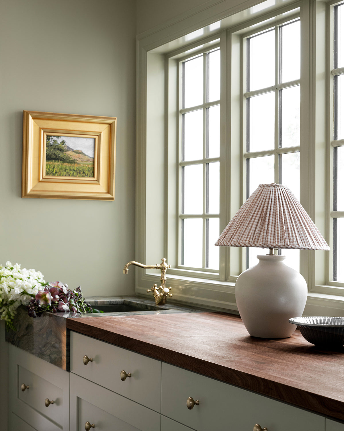 A kitchen with pale green cabinets, brass hardware, and a wood countertop features the Bronwyn Table Lamp with a gingham pleated shade, fresh flowers, and a sink under large windows. A framed landscape painting decorates the wall by McGee & Co.