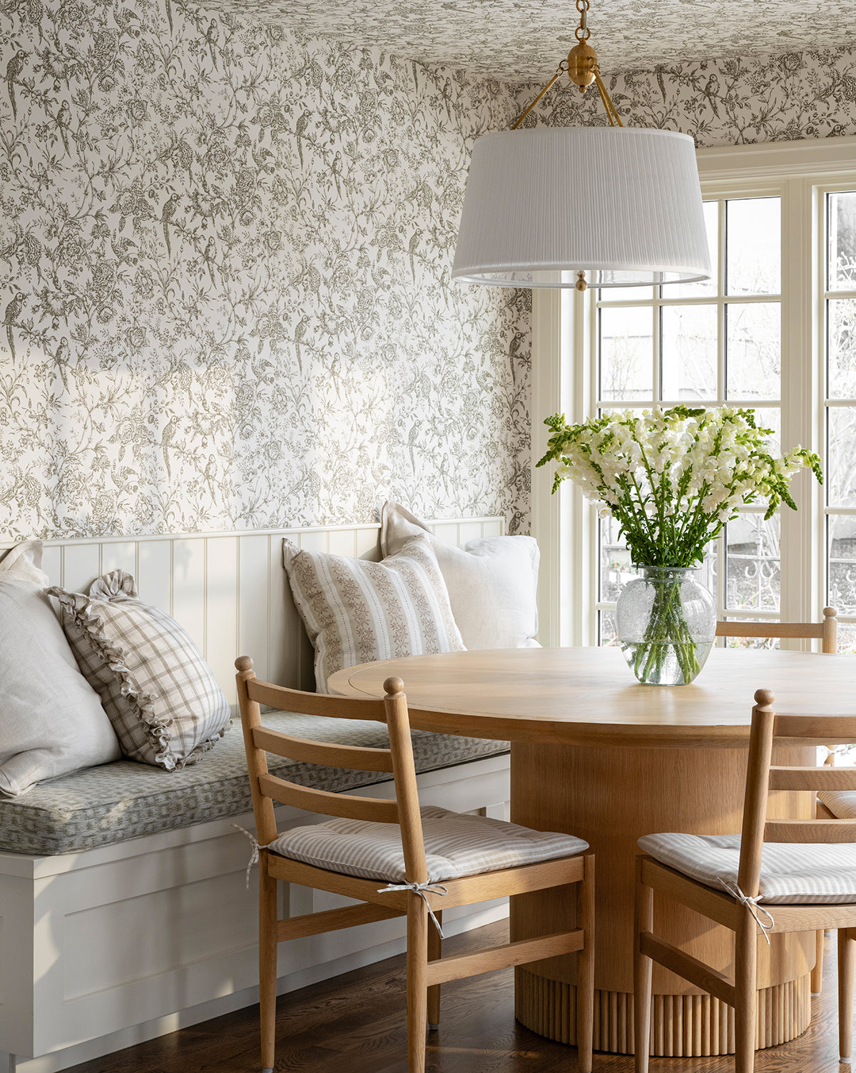 A cozy breakfast nook features the Gilroy Round Oak Dining Table with a reeded base in heritage oak, wooden chairs with cushions, a built-in bench with pillows, floral wallpaper, and large windows that fill the space with natural light by McGee & Co.