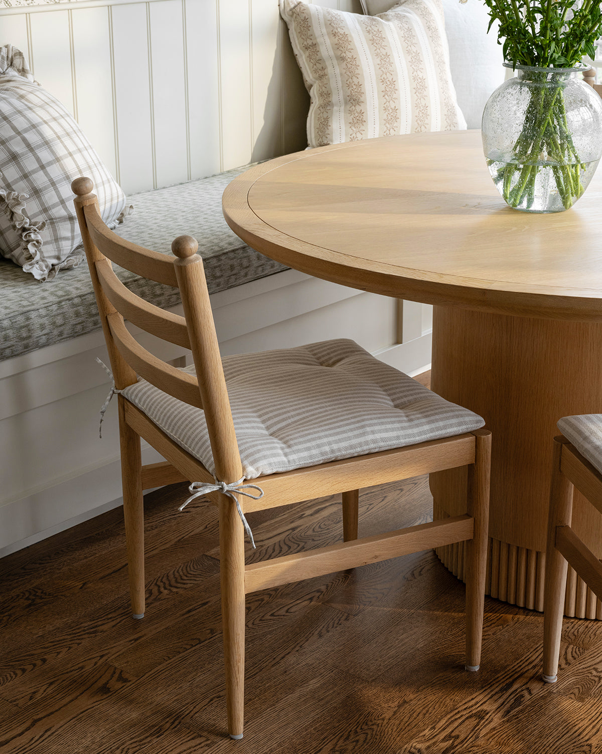 The Romany Dining Chair with a linen cushion sits by a round wooden table. Nearby, a bench with plaid pillows and a glass vase of greenery rest on wood floors in a cozy, sunlit dining nook, by McGee & Co.