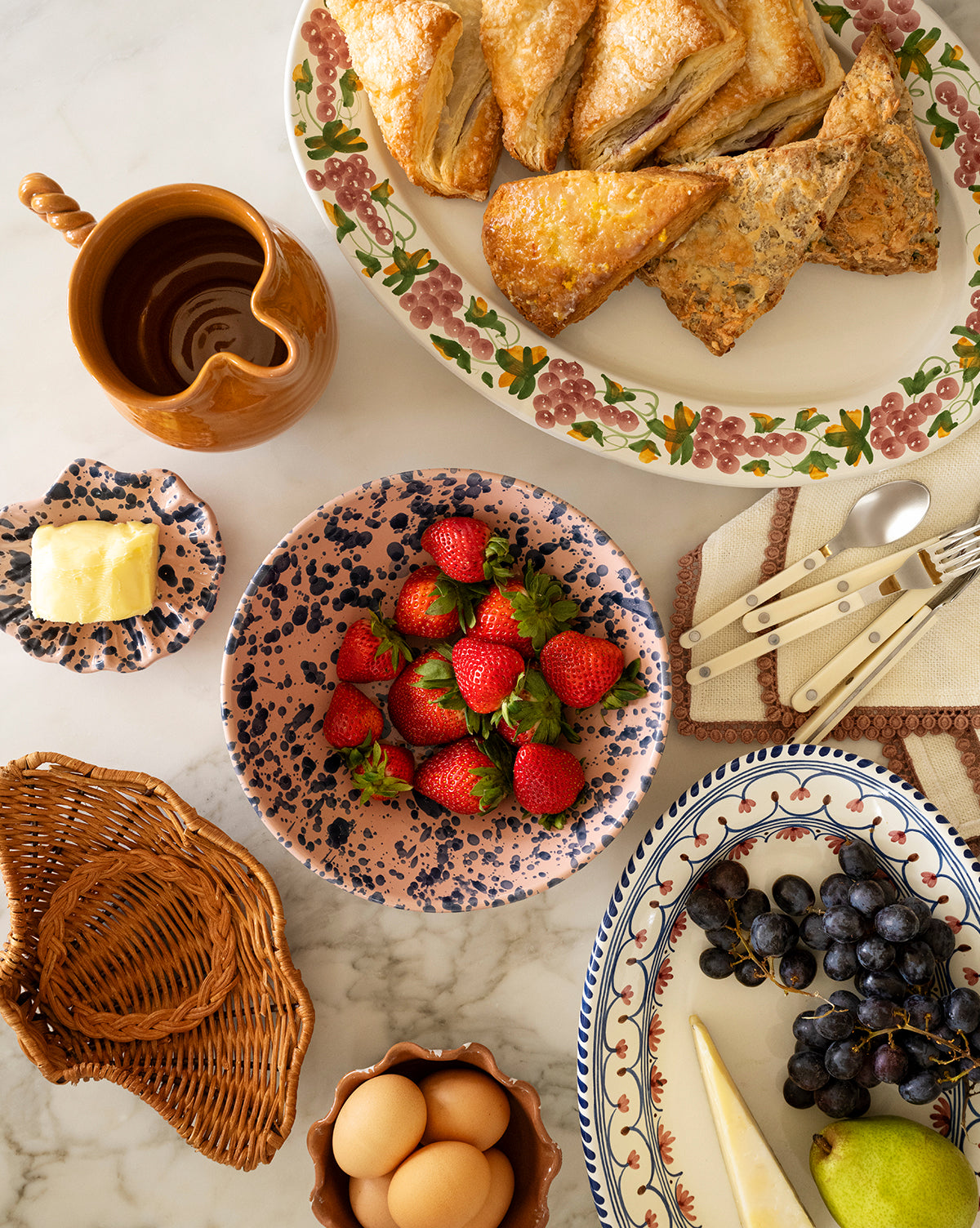 A breakfast table features a floral platter of pastries, fresh fruit, a small dish of butter, the rustic brown Louise Jug with a twisted handle, eggs in a basket, and utensils on a cloth napkin, by McGee & Co.