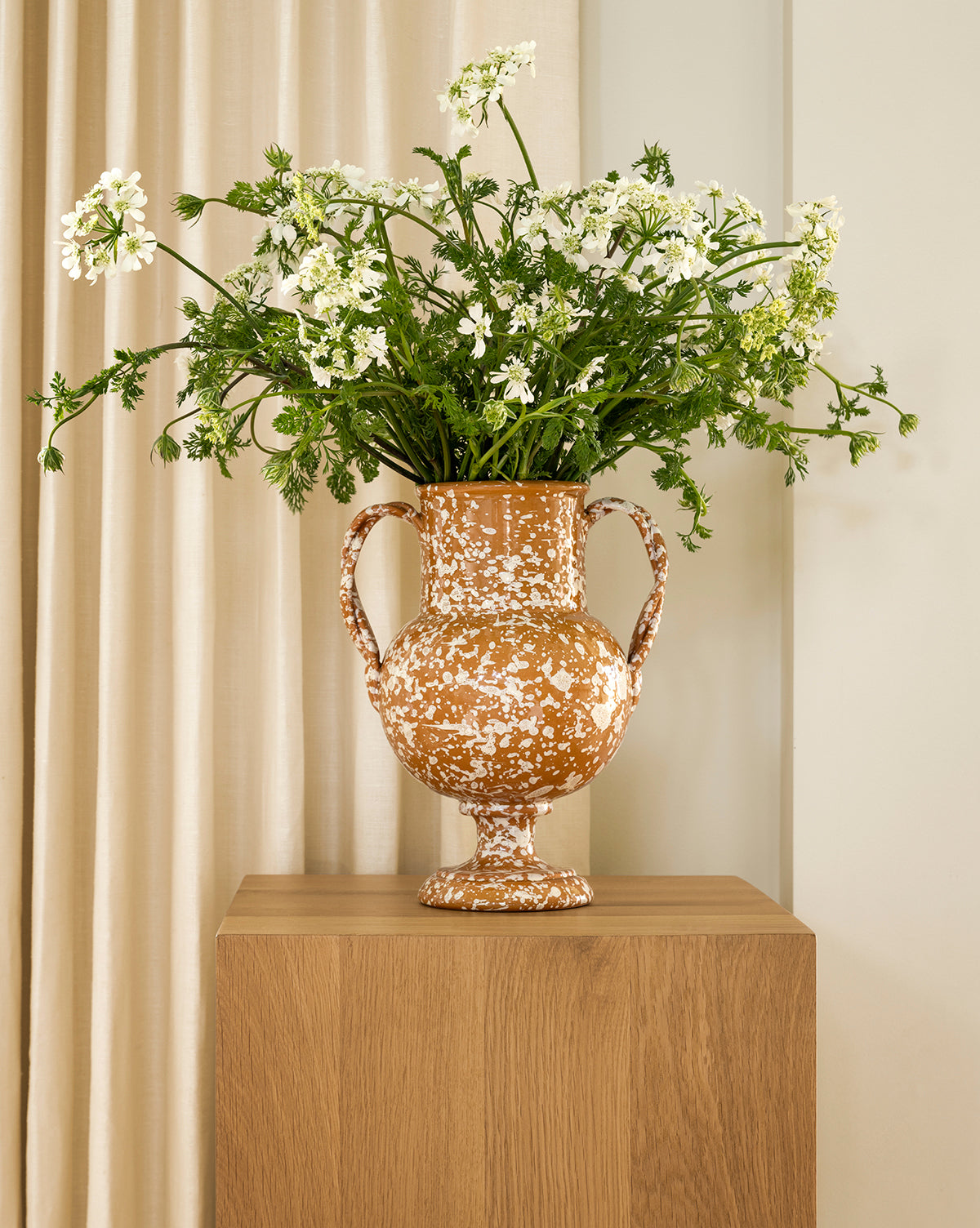 The Splatter Verona Vase, featuring two handles and a speckled design, holds white flowers and green foliage on a wooden pedestal against neutral beige curtains, by McGee & Co.