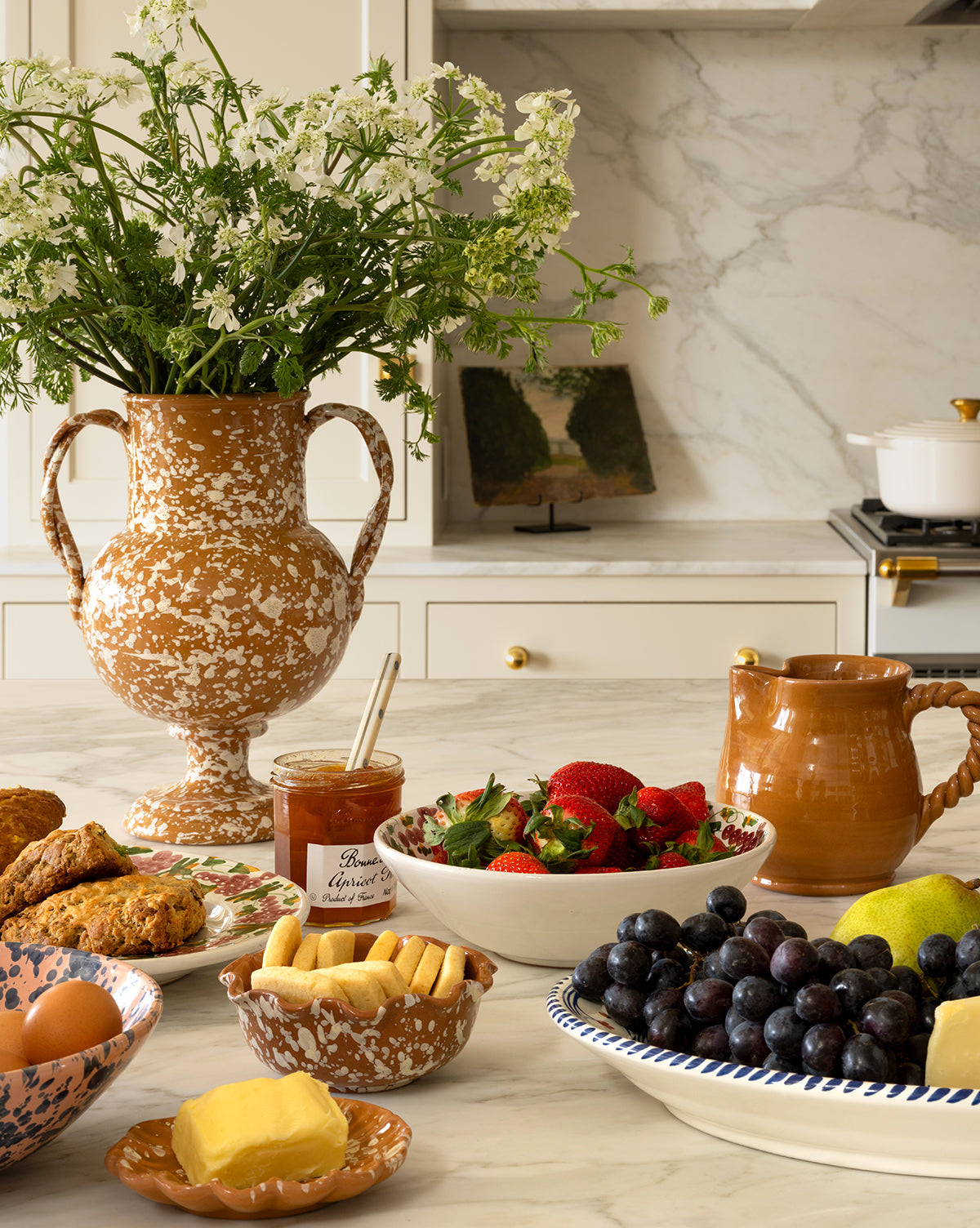 A kitchen counter displays the Splatter Verona Vase filled with white wildflowers, surrounded by strawberries, grapes, jam, pottery mugs, scones, butter, eggs, pears, and cookies—all beautifully arranged for breakfast, by McGee & Co.