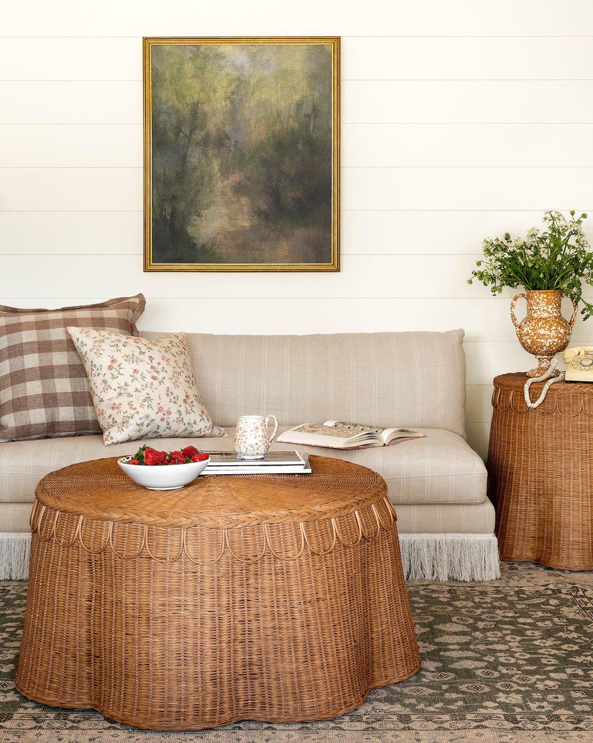 A cozy living room features a beige striped sofa, floral and plaid pillows, the Sharland England LTD Adeline Coffee Table with strawberries and a mug, a side table with flowers, and a framed painting on a white shiplap wall—a space curated in the inviting style of McGee & Co.