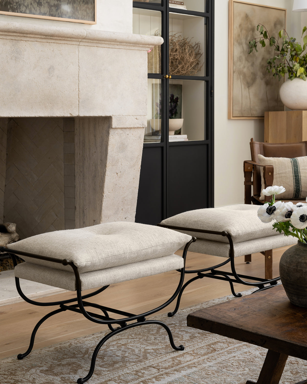 Two Makerspalm Alameda Stools with linen upholstery and matte black iron bases are placed in front of a stone fireplace in a cozy living room, accompanied by a wooden coffee table, vase of flowers, and decorative shelves. McGee & Co.