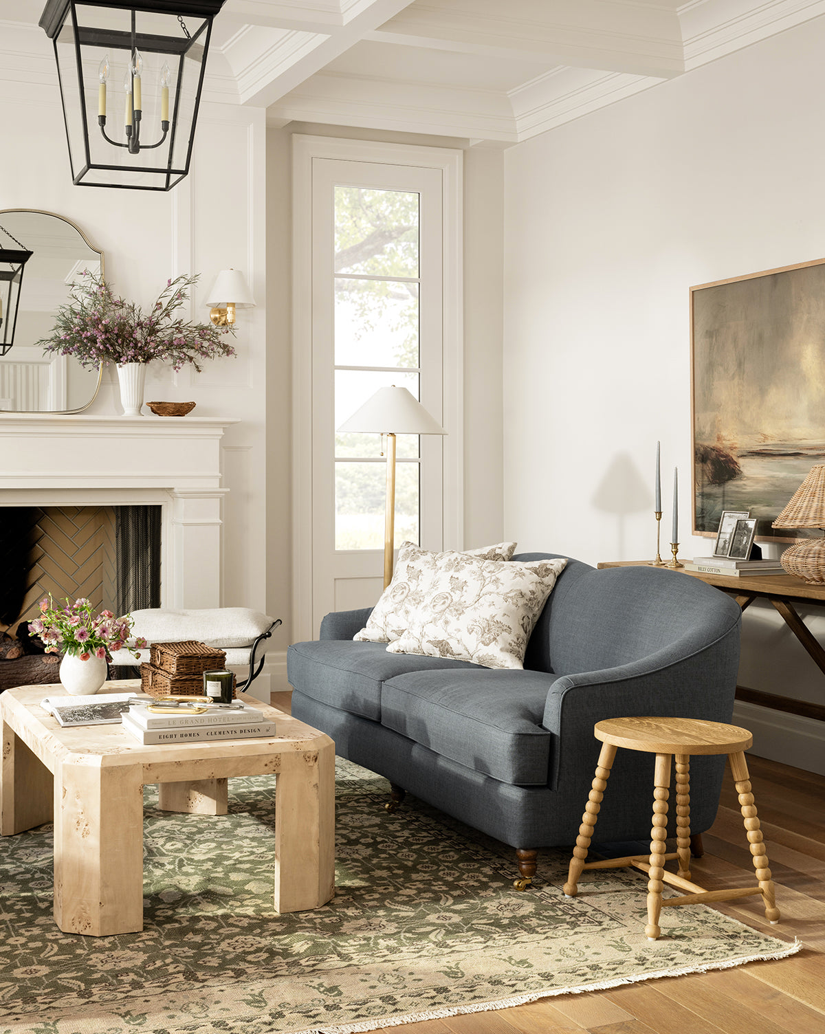 A cozy living room with a blue sofa, floral pillows, wooden coffee table, and the Fernwood Hand-Knotted Wool Rug in green tones. A fireplace topped with flowers and large windows complete the scene alongside decorative accents and artwork by McGee & Co.