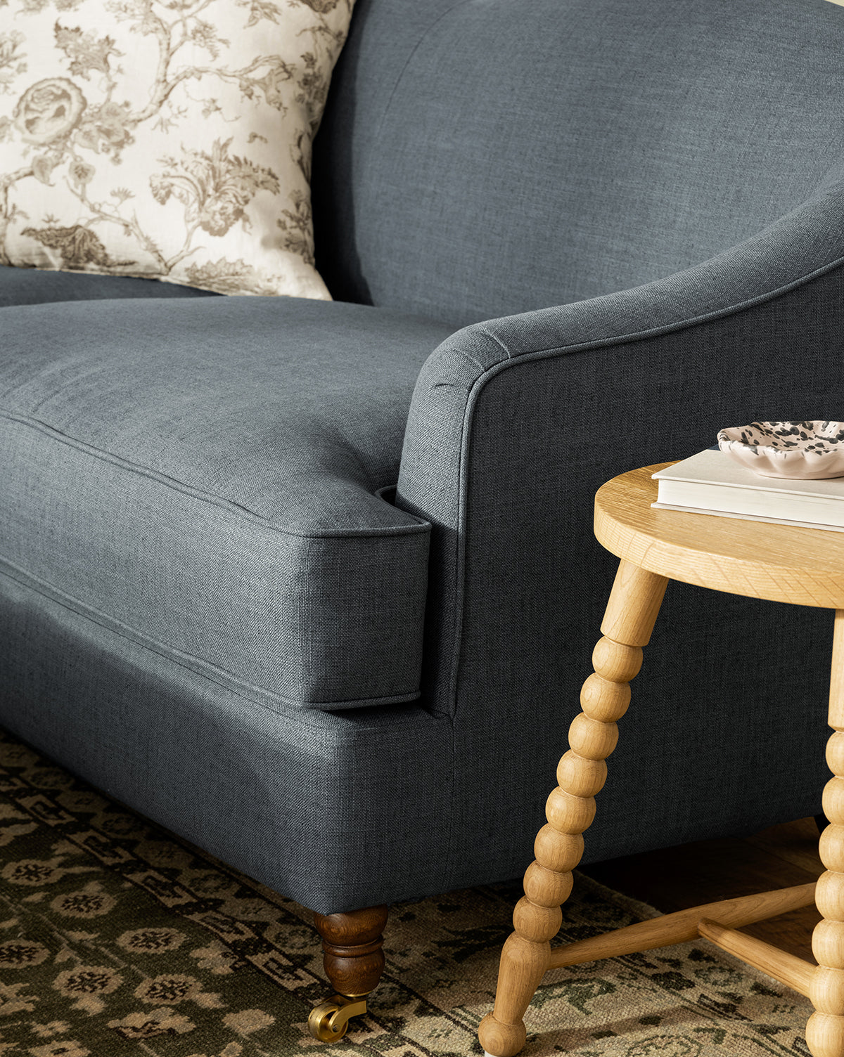 A close-up of the Fletcher Sofa 76 in dark gray, styled with a floral pillow beside a round wooden side table holding a book and decorative dish, sits atop a patterned rug—perfect for adding handcrafted style and comfort to your space, by McGee & Co.