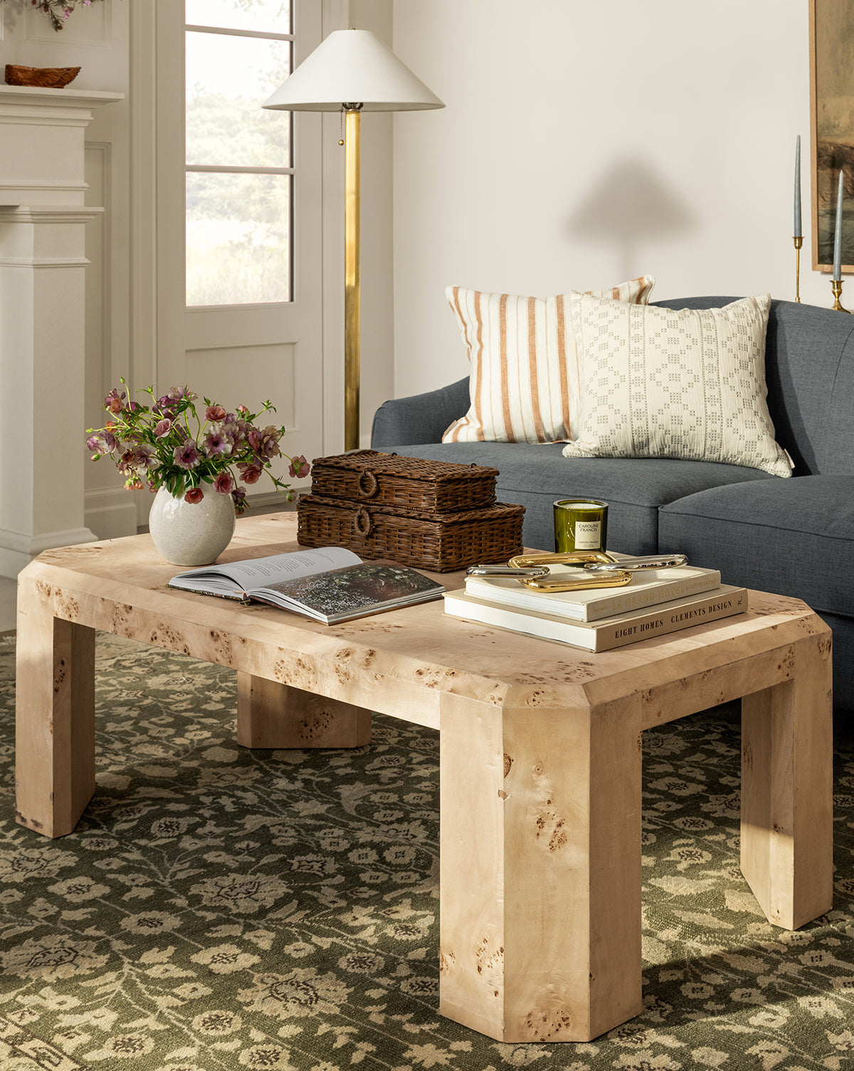 The Rocio Burl Coffee Table holds books, a vase of flowers, a candle, and wicker boxes in a cozy living room with a blue sofa, patterned cushions, a floor lamp, and a patterned rug covering the floor, by McGee & Co.