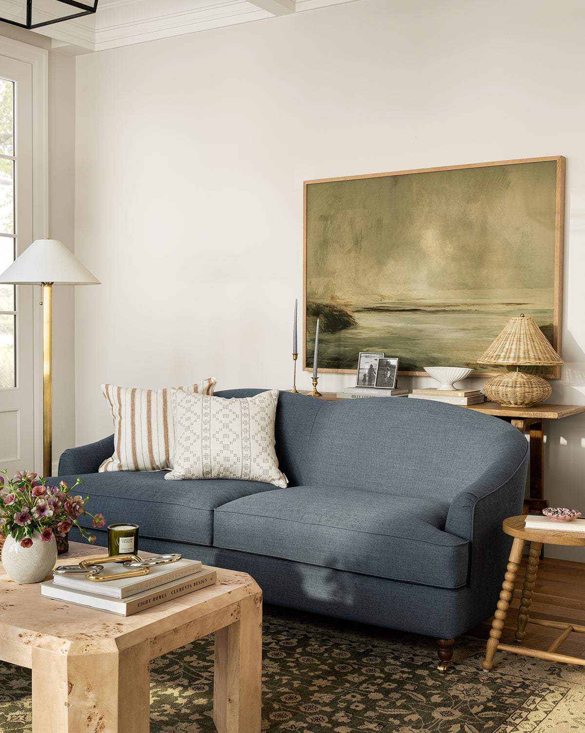 A cozy living room with a blue sofa, patterned pillows, a wood side table and woven lamp, a tall floor lamp, and the Misty Blue Headland gallery-wrapped landscape canvas above a coffee table styled with books and flowers, by McGee & Co.