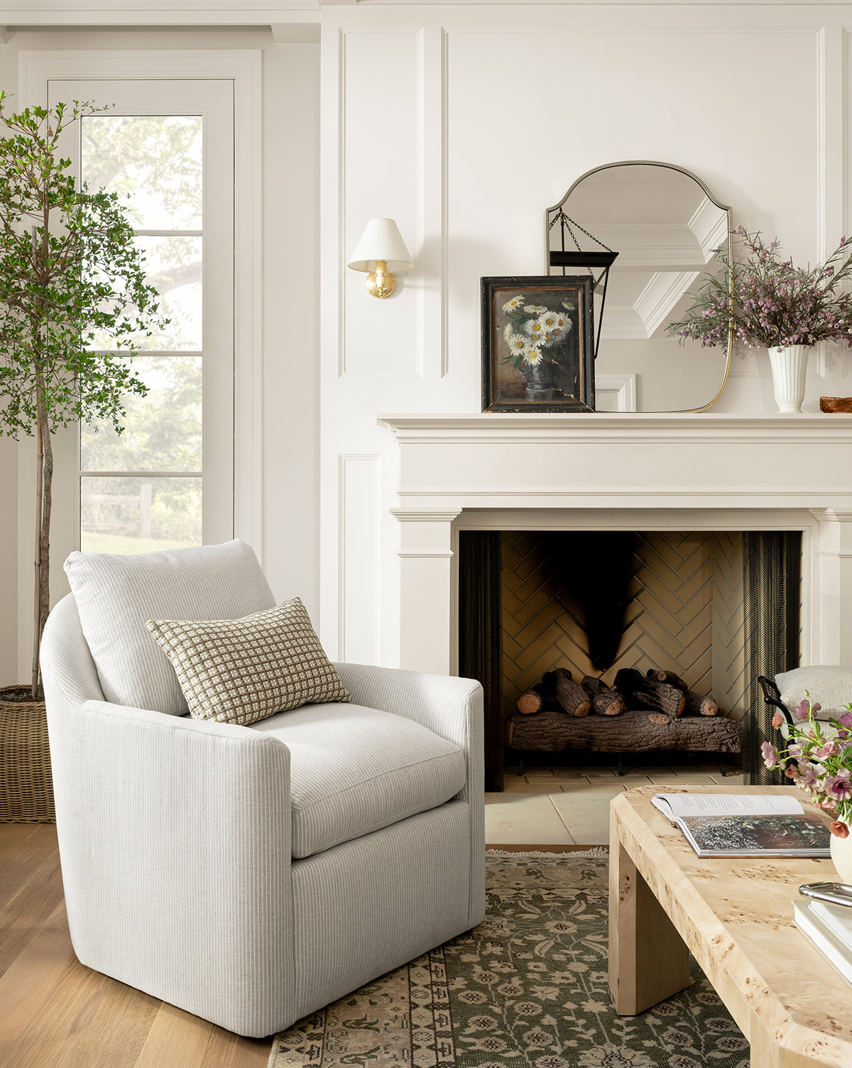A cozy living room features a light gray armchair with a patterned pillow, wooden coffee table, fireplace with logs, Sussex Wall Mirror above the mantel, artwork, flowers, and a window overlooking greenery by McGee & Co.
