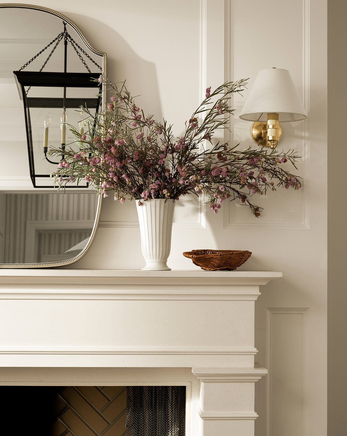 A white mantel displays the Delphi Footed Vase filled with pink and green flowers next to a small woven basket. Above, a large mirror, lantern-style light fixture, and brass wall sconce hang on the cream-paneled wall, by McGee & Co.
