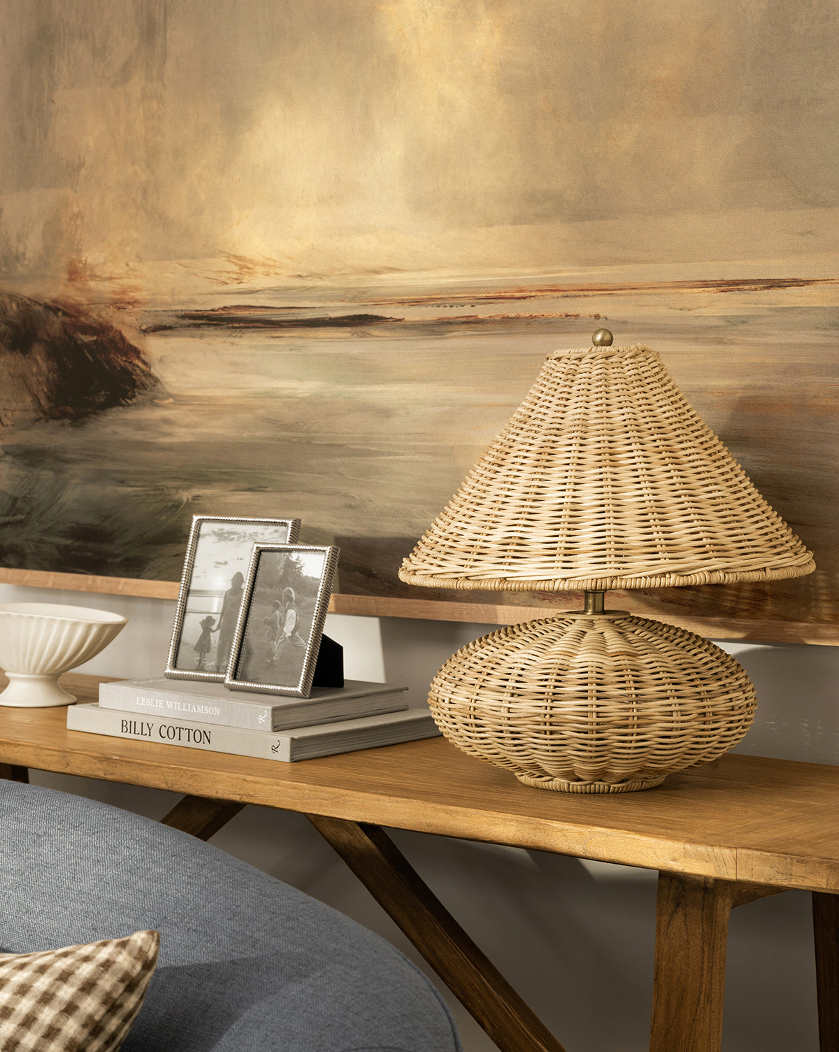 The Palmetto Table Lamp with a woven shade sits on a wooden console table beside framed black-and-white photos, a white bowl, and books, with a large landscape painting behind—perfect for organic home decor, by McGee & Co.