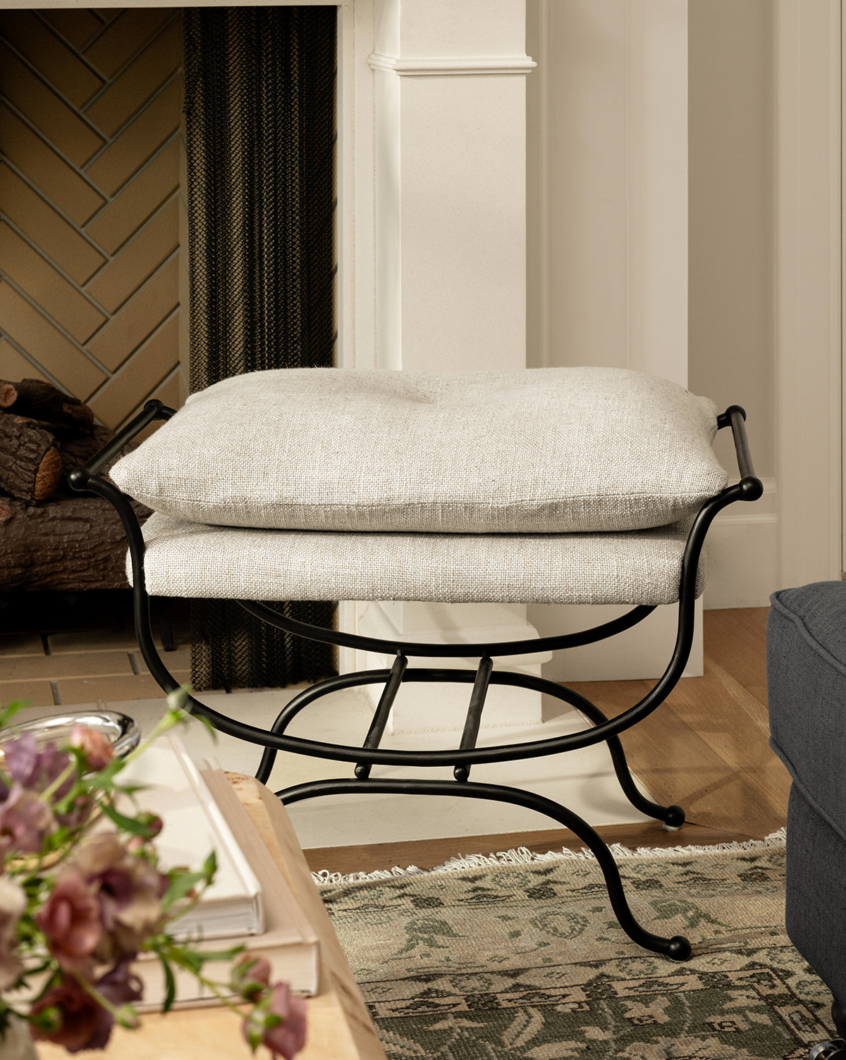 A cozy living room showcases the Makerspalm Alameda Stool with a matte black iron base and linen upholstery by the fireplace, next to a blue sofa and wooden table with flowers on a patterned rug, McGee & Co.