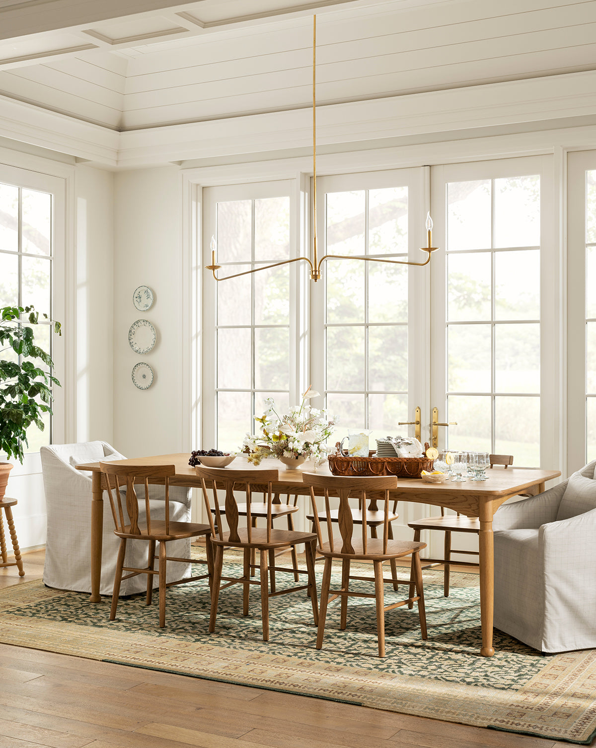 A bright dining room features the handcrafted Phipps Dining Table set for a meal, six wooden chairs, two white armchairs, a brass modern chandelier, large windows, wall plates, a potted plant, and a patterned rug on wooden floors, by McGee & Co.