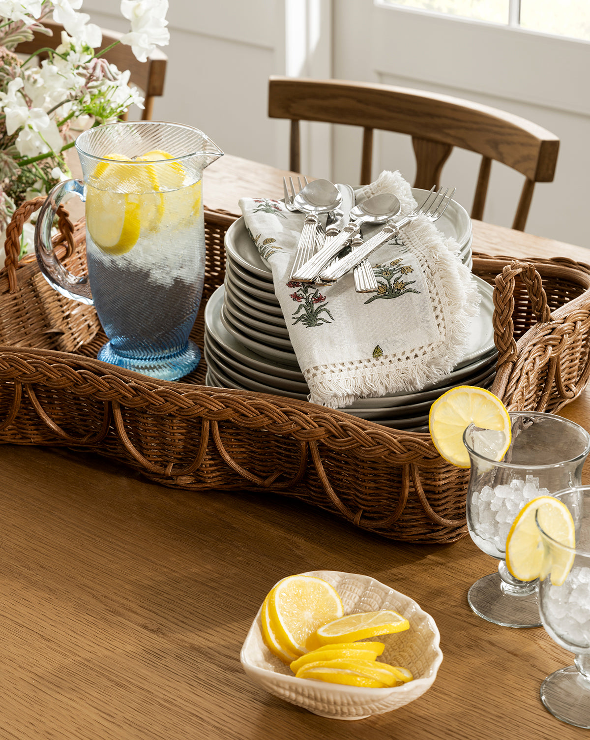 A wicker tray holds the Spiral Jug filled with lemon water, stacked plates, and cutlery with napkins. Handcrafted glass tumblers with lemon slices and a bowl of lemon wedges sit nearby, all bathed in bright natural light by McGee & Co.