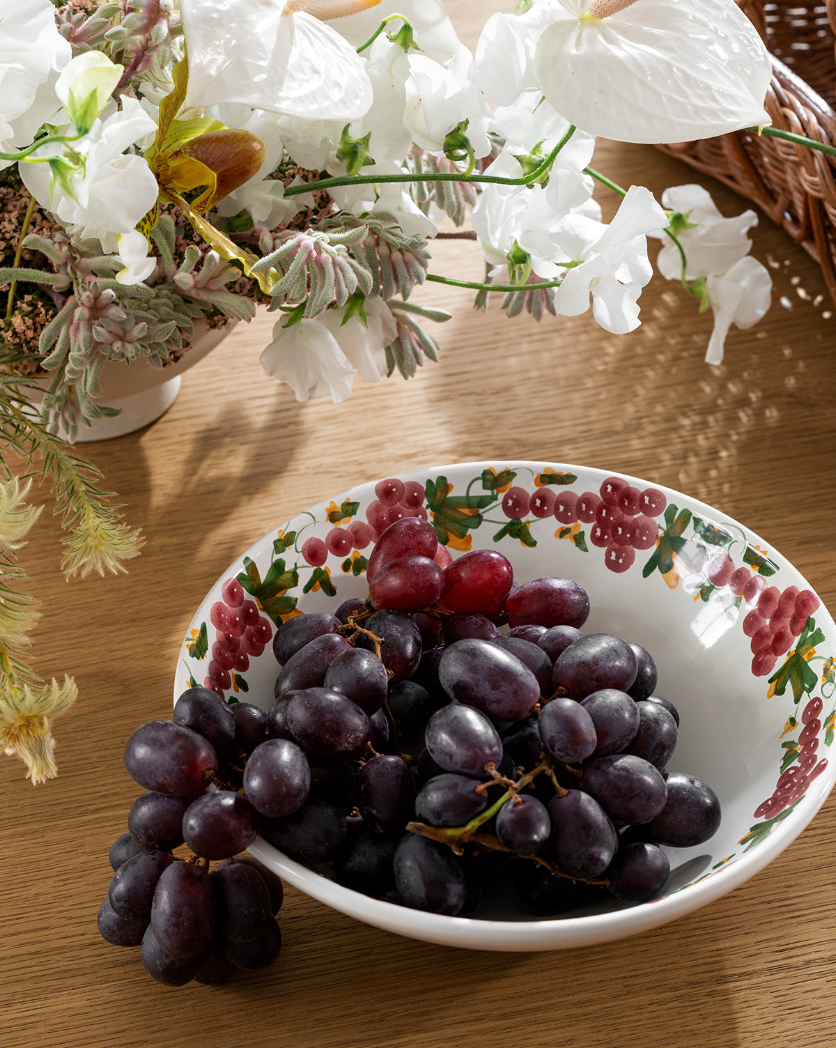 The Vine Large Bowl, a handmade ceramic bowl with a grapevine motif, holds bunches of purple grapes on a wooden table. A white floral arrangement and part of a wicker basket add to the charming scene by McGee & Co.