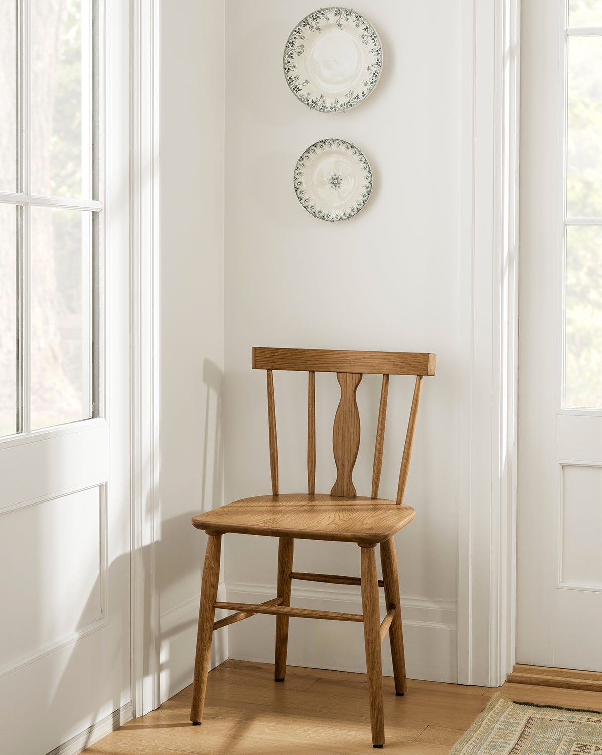 A wooden dining chair with a spindle back design, placed in a corner of a room with a light interior, decorated with framed decorative plates on the wall in the background.