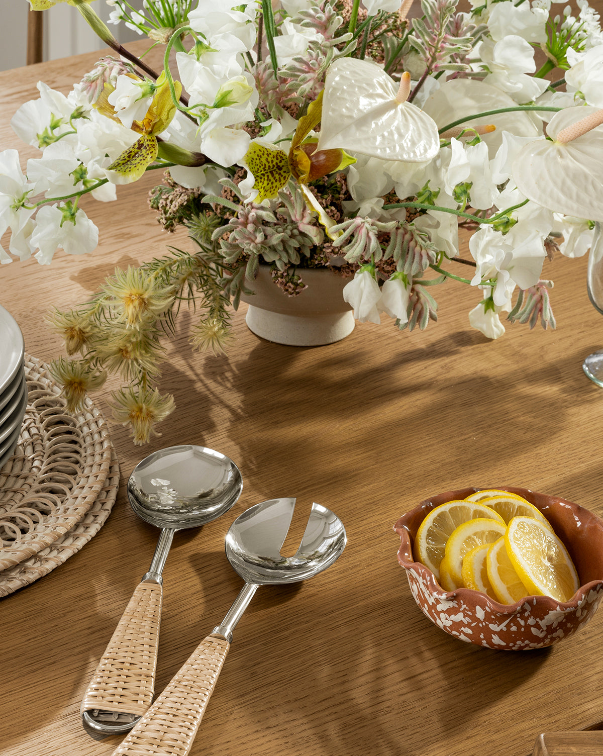 A wooden table with a ceramic vase of white flowers, two woven-handled serving utensils, a basket of stacked plates, and the Splatter Scallop Dish filled with lemon slices, by McGee & Co.