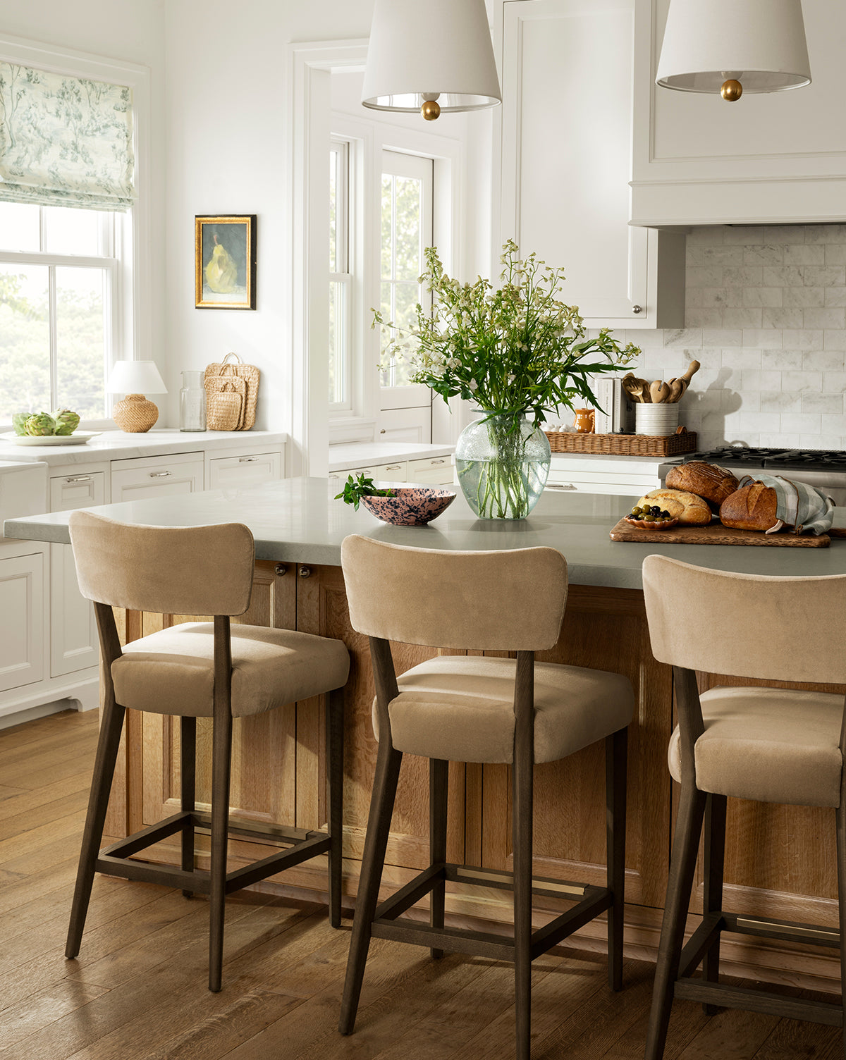 Bright kitchen with a wooden island, three Makerspalm Etienne Stools (Ready to Ship), glass vase with flowers, bread and fruit on the counter, white cabinets, and large windows letting in natural light, McGee & Co.
