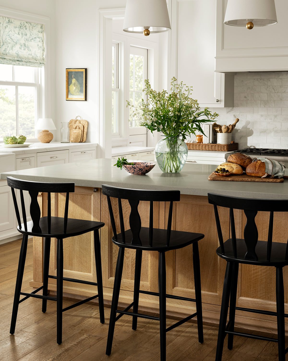 A bright, modern kitchen with a light wood island features three Whittaker Counter Stools by YANG CHENG WOODEN INDUSTRIES INTERNATIONAL (VIETNAM) CO LTD, each with a spindle back, plus fresh bread and flowers on the counter for an airy vibe—finished with design touches inspired by McGee & Co.