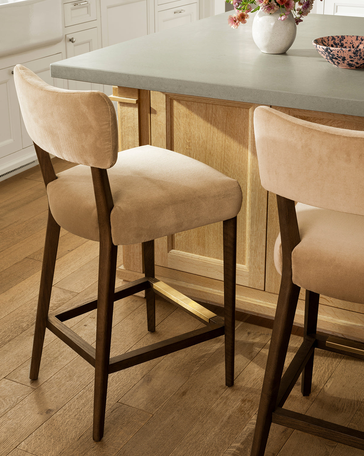 Two beige upholstered Makerspalm Etienne Stools (Ready to Ship) with dark wood legs sit by a light wood kitchen island with a gray countertop, where a vase and bowl accentuate the customizable stool design and complement the light wood floor. McGee & Co.