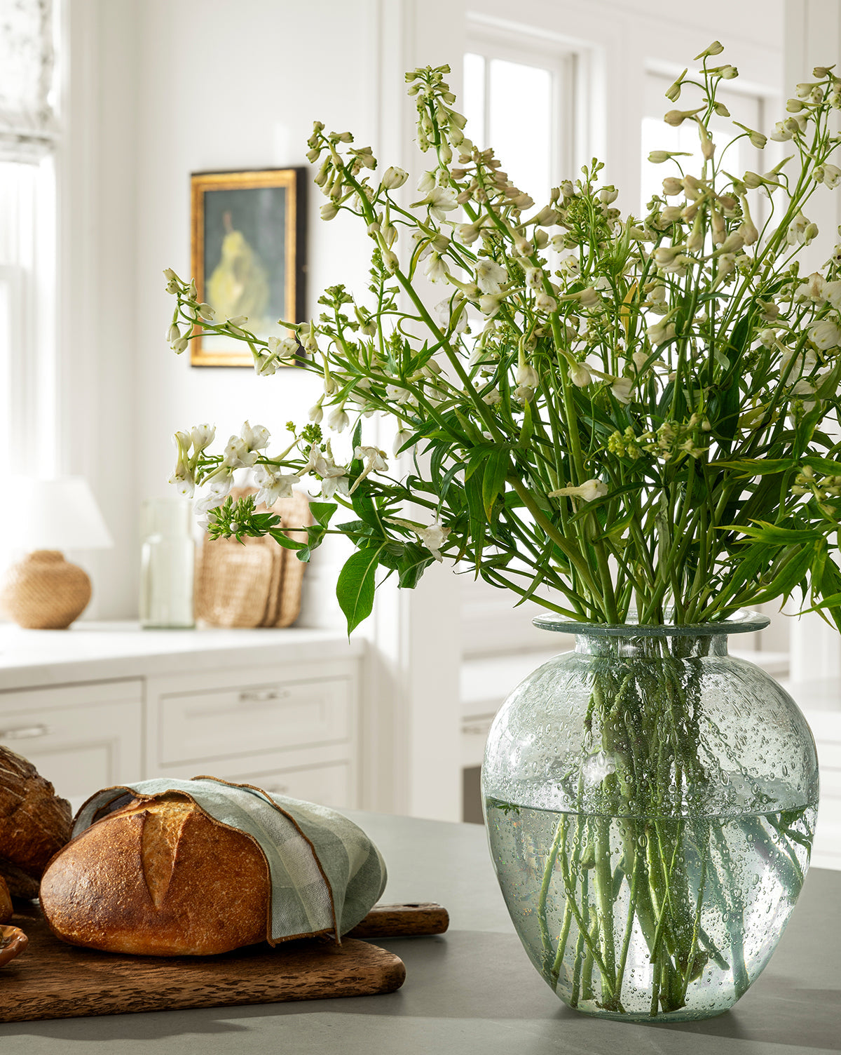 The Gretchen Vase, filled with tall white flowers, sits on a kitchen counter beside a loaf of bread on a wooden board with a linen cloth. The bright kitchen showcases white cabinets and natural decor, by McGee & Co.