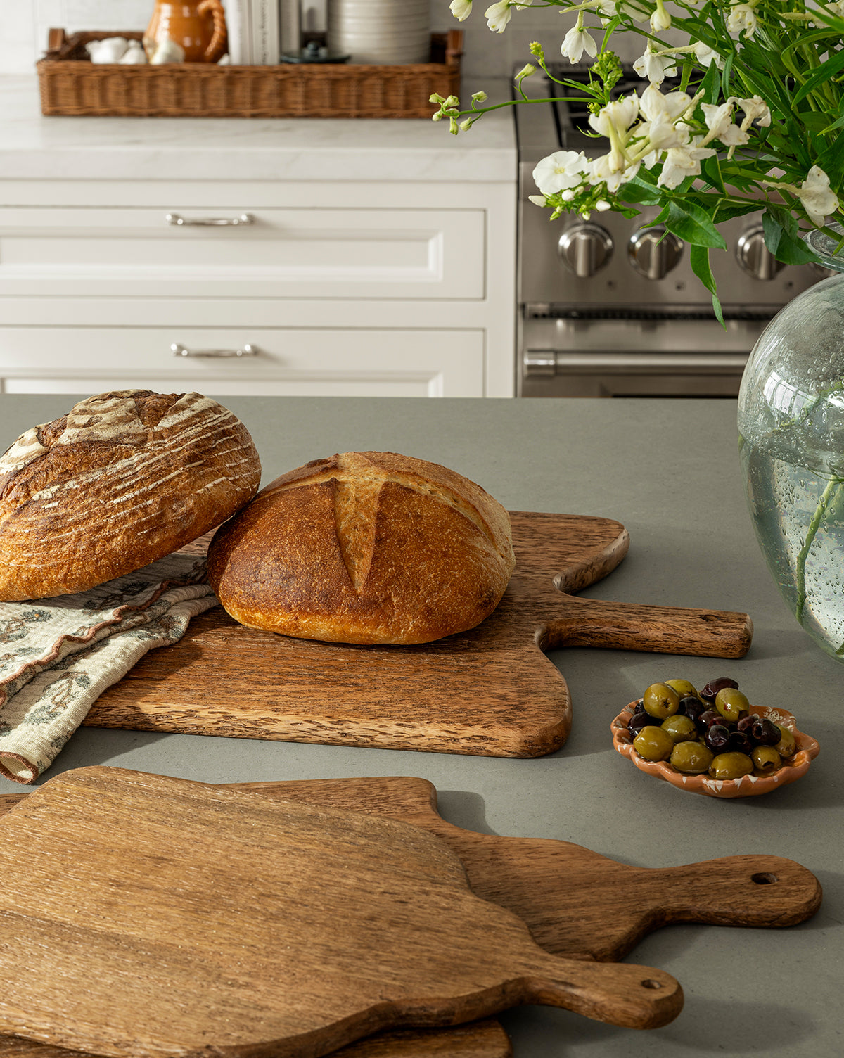 Two round loaves of bread rest on Longfellow Breadboards on a kitchen counter next to a small dish of olives. White cabinets and a vase of flowers add to the cozy kitchen decor in the background, by McGee & Co.