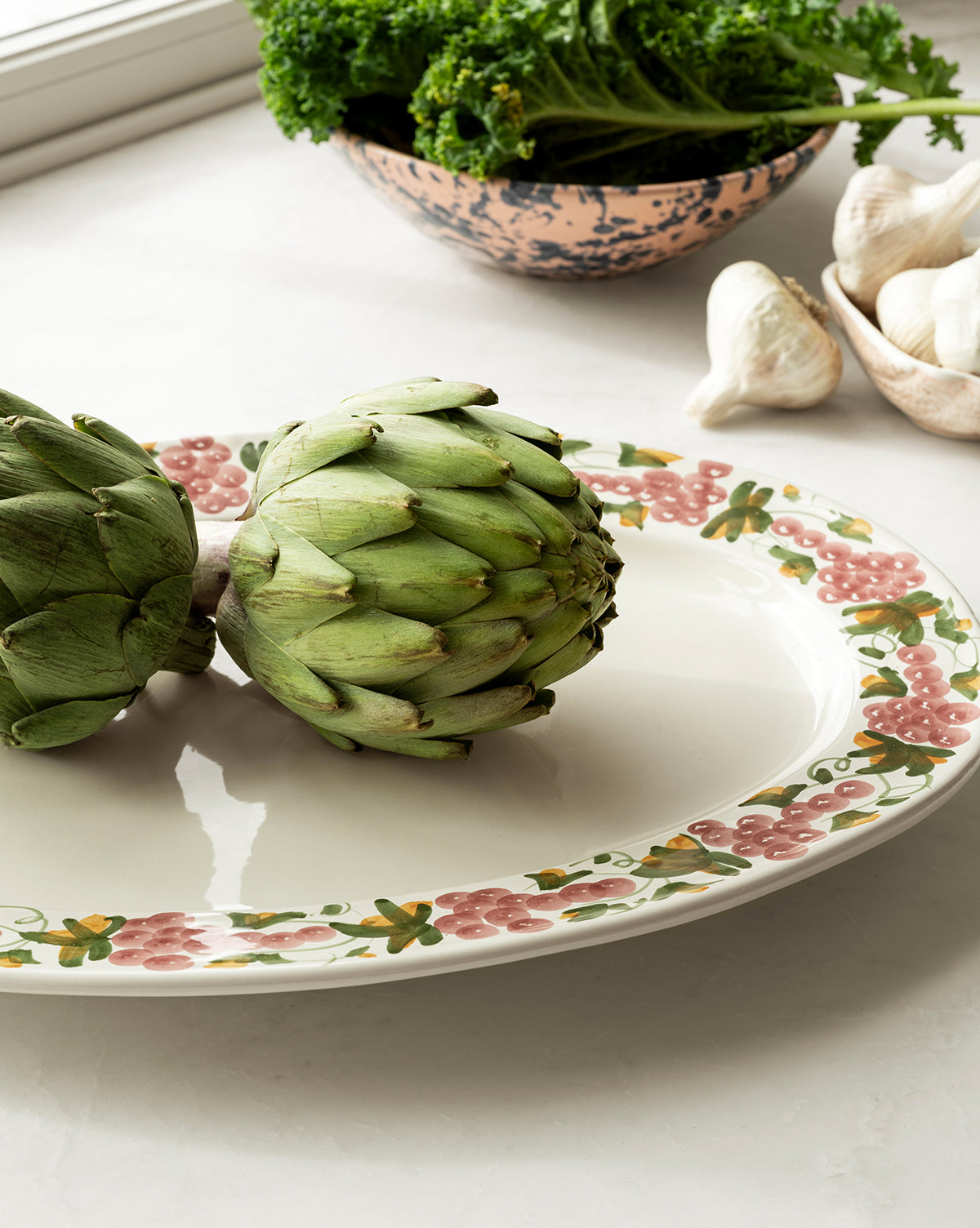 Two fresh artichokes sit on the Vine Serving Platter, which features a floral border; in the background are leafy greens in a bowl and a garlic bulb on a white countertop near a window, by McGee & Co.