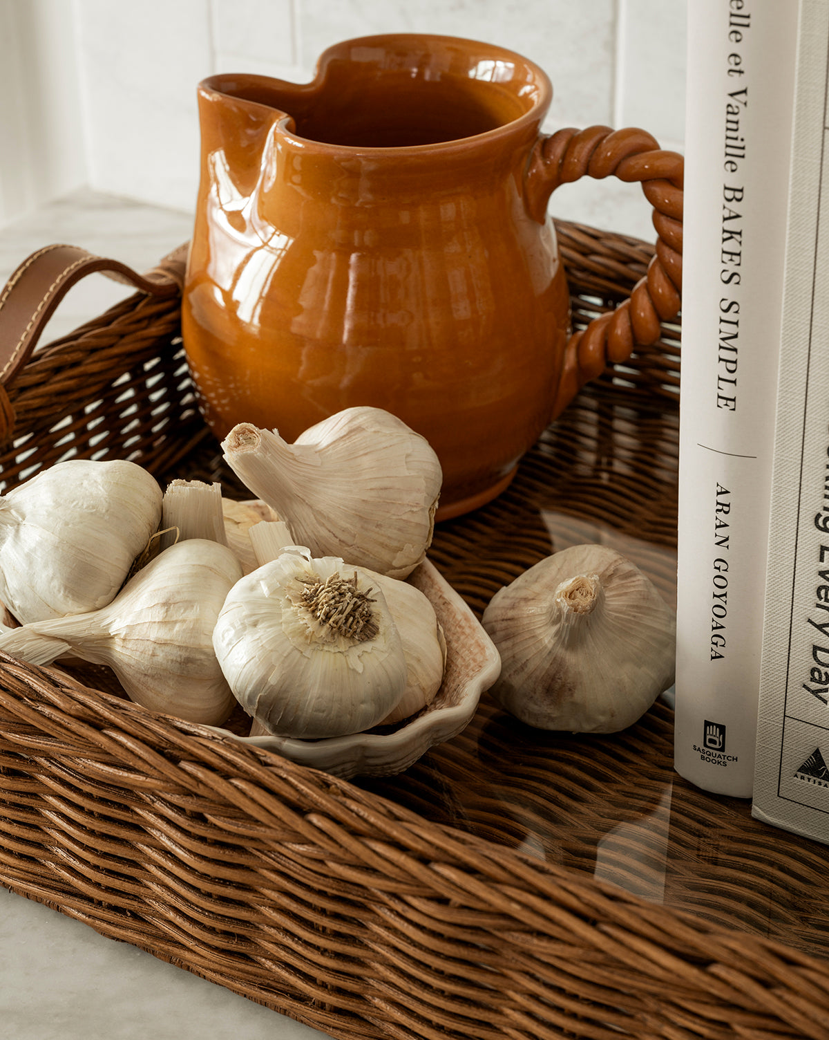 The Ashcroft Wicker Tray displays garlic bulbs in a shell-shaped dish, an orange ceramic pitcher with a twisted handle, and upright white cookbooks on a light glass countertop, by McGee & Co.