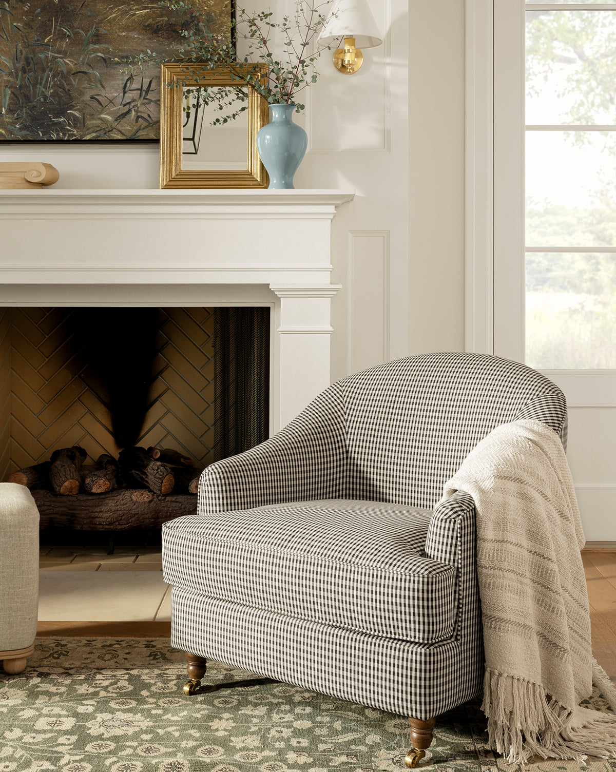The Makerspalm Fletcher Lounge Chair, featuring a black-and-white checkered pattern, sits by the fireplace with a textured cream throw draped over one arm. Above the mantel are a vase, small frame, and nature painting by McGee & Co.