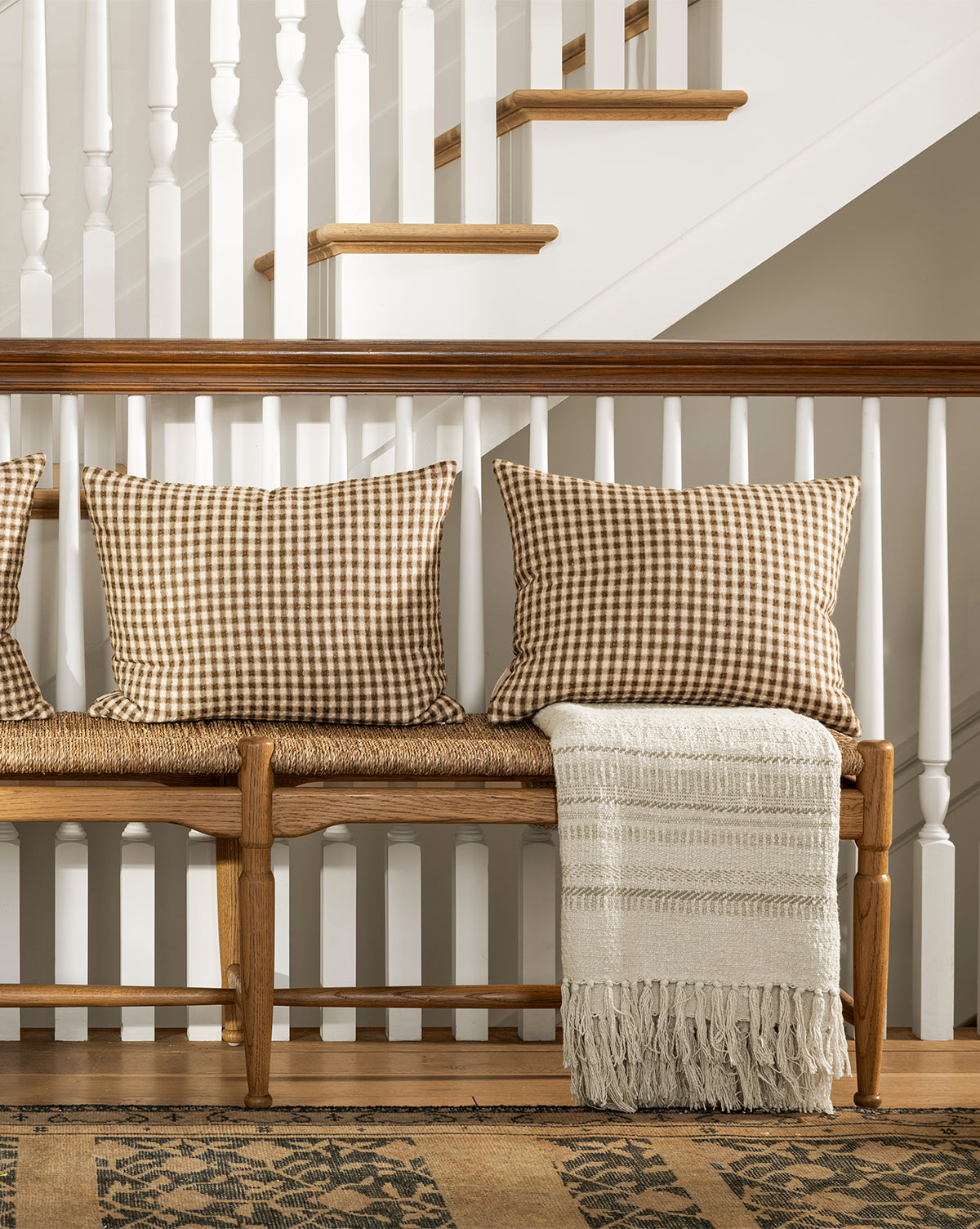A wooden bench with two brown gingham pillows and the Donahue Striped Throw by ILA HOME FASHIONS sits against a staircase with white spindles and wood accents, with a patterned rug on the floor below, styled by Studio McGee & Co.