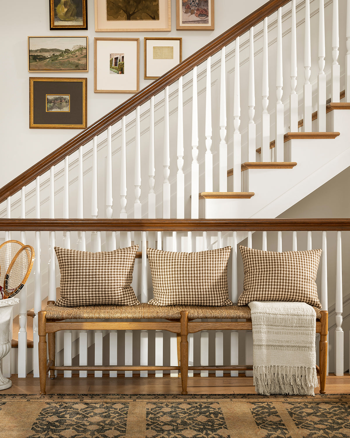 A cozy hallway features the Phipps Woven Bench topped with three plaid pillows and a fringed throw, set by a white staircase with art. A patterned rug and small table display vintage tennis rackets, while honey oak tones warm the space, by McGee & Co.