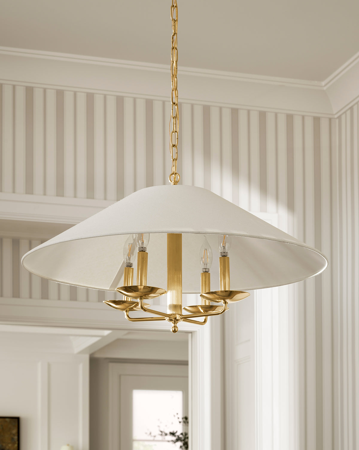 The Townes Chandelier by Meredith (China) Ltd features a large linen shade, gold metal frame, and four candle-style bulbs, hanging from a gold chain in a room with striped wallpaper and white trim—McGee & Co.
