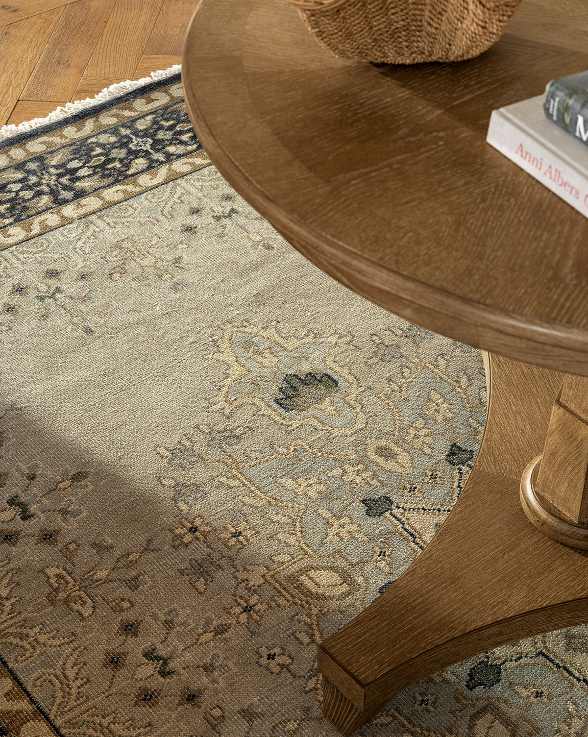 A close-up of a round wooden table on the Deveraux Hand-Knotted Wool Rug in beige and blue, with a woven basket and books on top. The table’s leg and the rug’s intricate design are clearly visible, by McGee & Co.