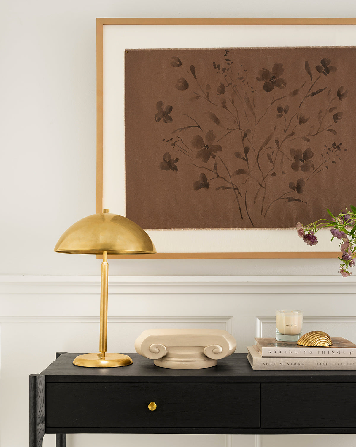The Aldorno Desk Table Lamp sits on a black console table alongside a decorative scroll, books, candle, and glass vase with flowers, beneath brown floral artwork. This display enhances the living rooms elegance against a white wall, by McGee & Co.
