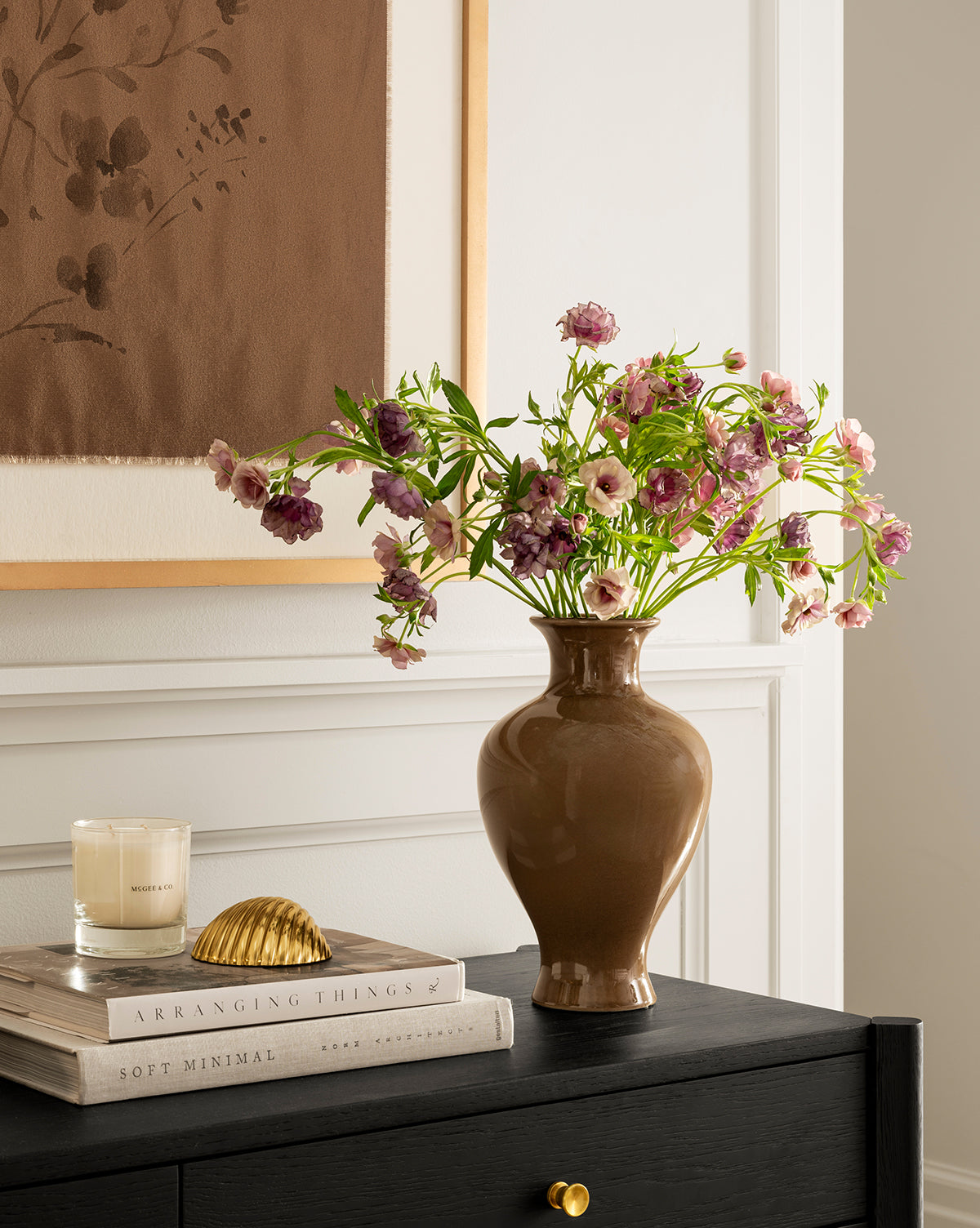 A brown Natalia Vase filled with pink and purple flowers sits on a black table next to a white candle, stacked books, and gold seashell decor, with framed artwork hanging on the wall in the background, by McGee & Co.