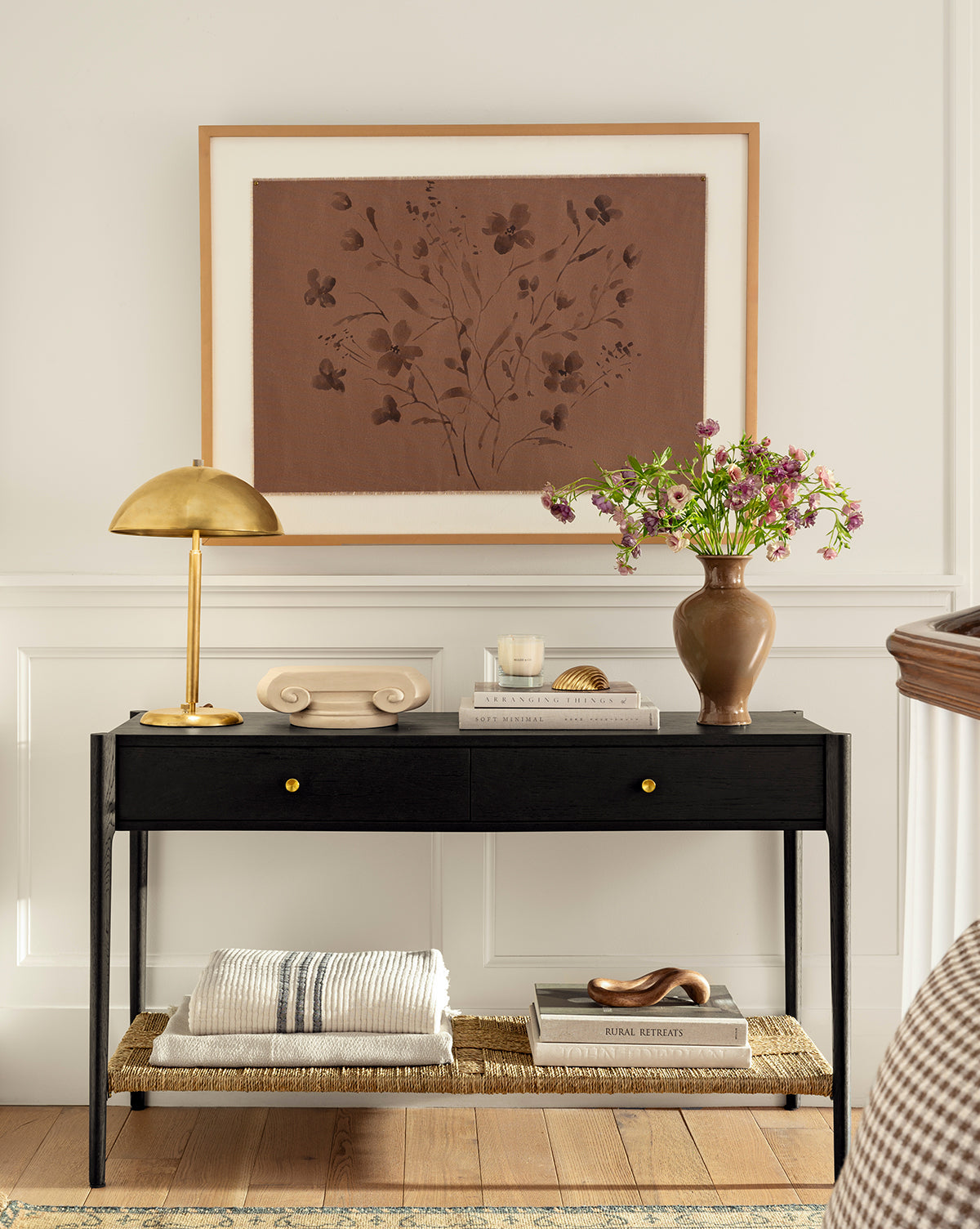 A Creative Co-op Anneliese Striped Throw is draped on a black console table with gold knobs, accented by books, a gold lamp, decor items, and a brown vase with pink flowers. Above hangs floral artwork in brown tones on a white paneled wall—ideal for luxury home decor. McGee & Co.