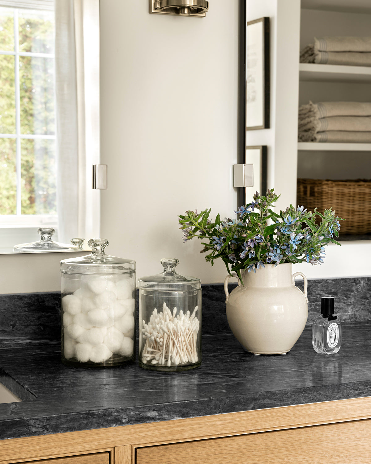 A Rasley Handled Jar sits on the bathroom counter beside glass jars of cotton balls and swabs, a cream vase with blue flowers, and a porcelain jar. Towels and a wicker basket are neatly stacked on shelves in the background, by McGee & Co.
