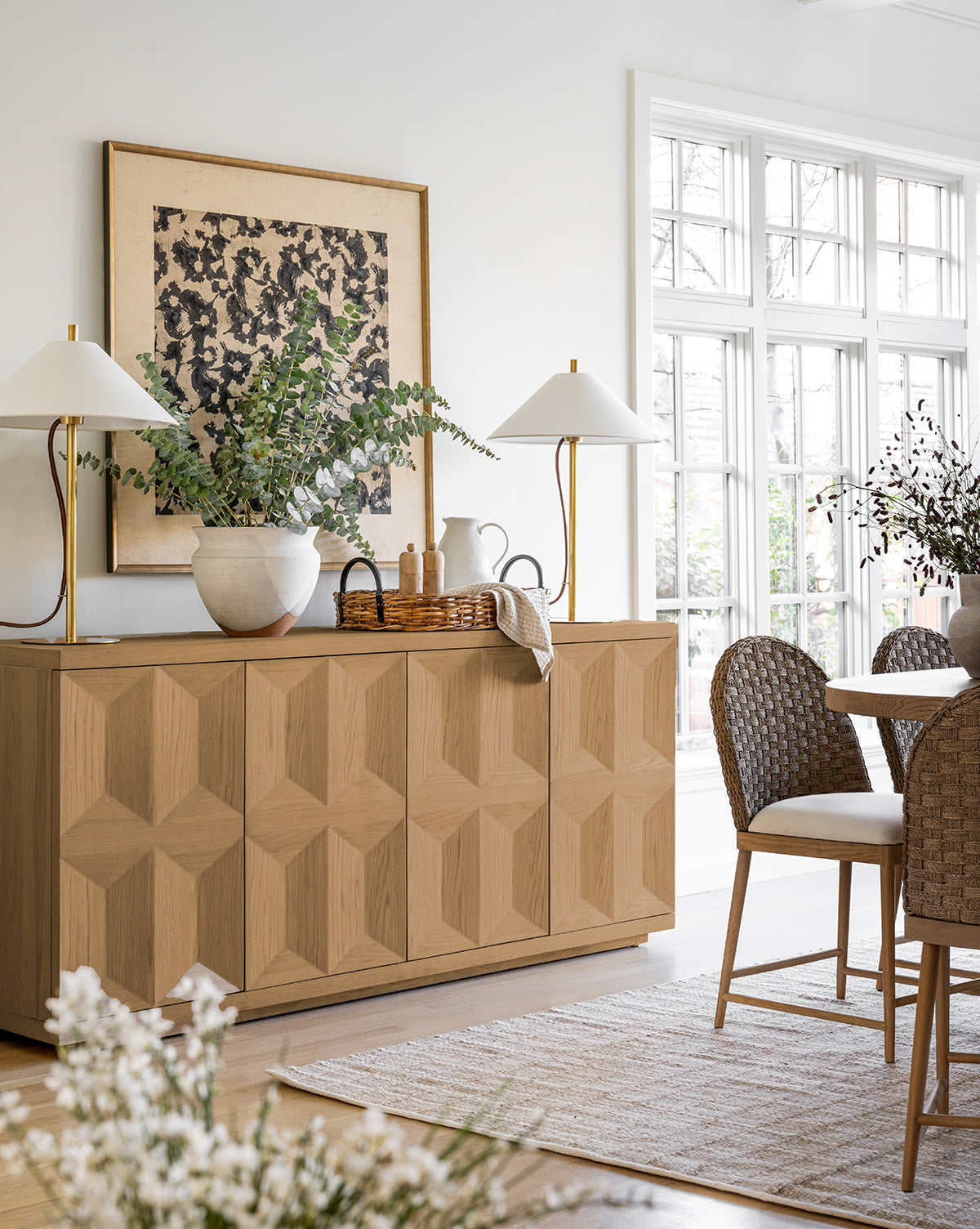 A bright dining room with woven chairs, the Beacon Sideboard featuring a geometric pattern and roomy storage, two white lamps, potted plants, large windows for natural light, and a framed abstract artwork above the cabinet, by McGee & Co.
