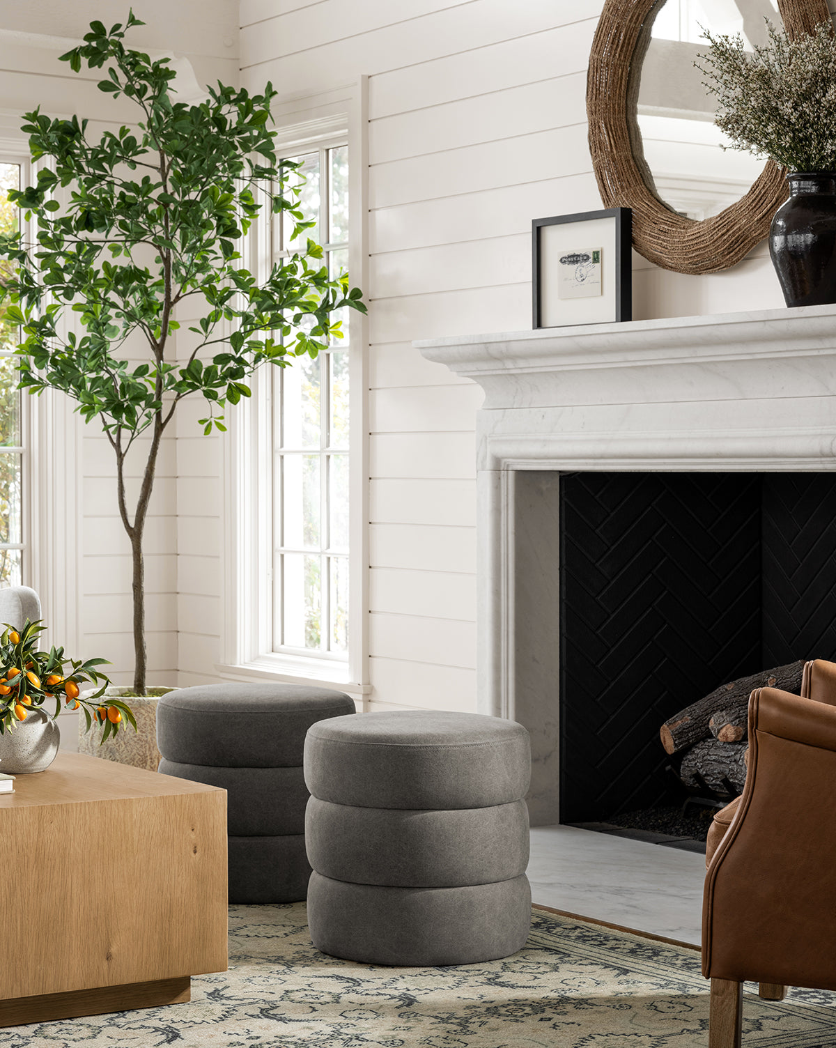 A cozy living room with a marble fireplace, two gray Channel Ottomans, a brown leather chair, a potted tree, a wooden coffee table with fruit, and large windows bringing in natural light for midcentury charm, by McGee & Co.