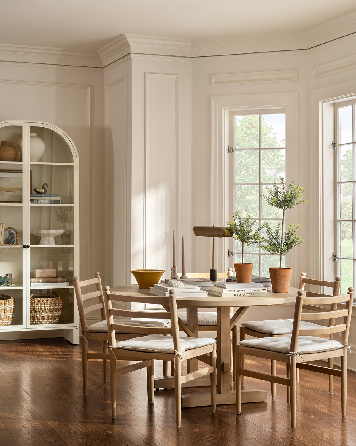 A bright, elegant dining room features a round white oak table, four Romany Dining Chairs with linen cushions, potted plants, books, and decorative objects. Large windows fill the space with light; a cabinet with shelves stands in the corner, by McGee & Co.