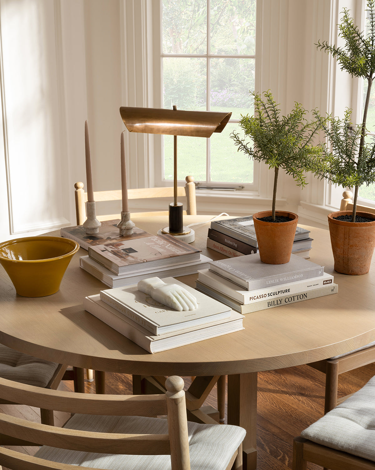 A round wooden table with stacked books, potted plants, a brass lamp, candles, the Lady Onyx Hand Object, and a yellow bowl sits by large windows. Wooden chairs on a hardwood floor complete the contemporary decor.