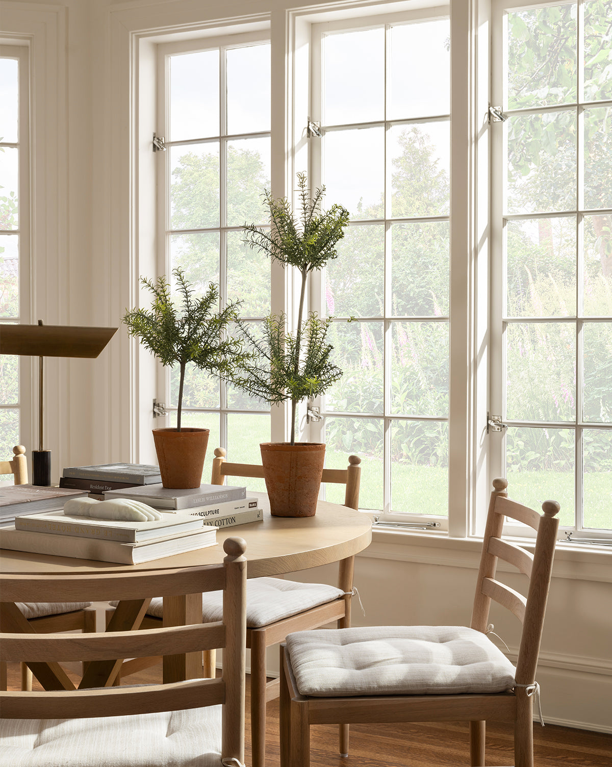 A sunlit dining area with a round wooden table and cushioned chairs features a Potted Faux Rosemary Topiary in a terracotta pot. Large windows overlook a green garden, filling the neutral-toned room with cozy natural light.