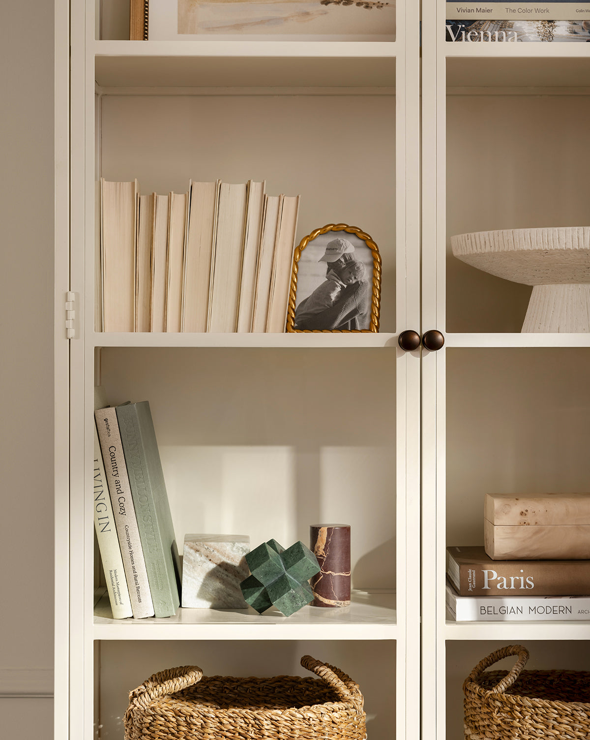 A beige bookshelf with books, art books, a framed B&W photo, the Diani Marble Cuboid Object as sculptural decor, and wicker baskets. The minimalist arrangement creates an organized, contemporary look for your home.